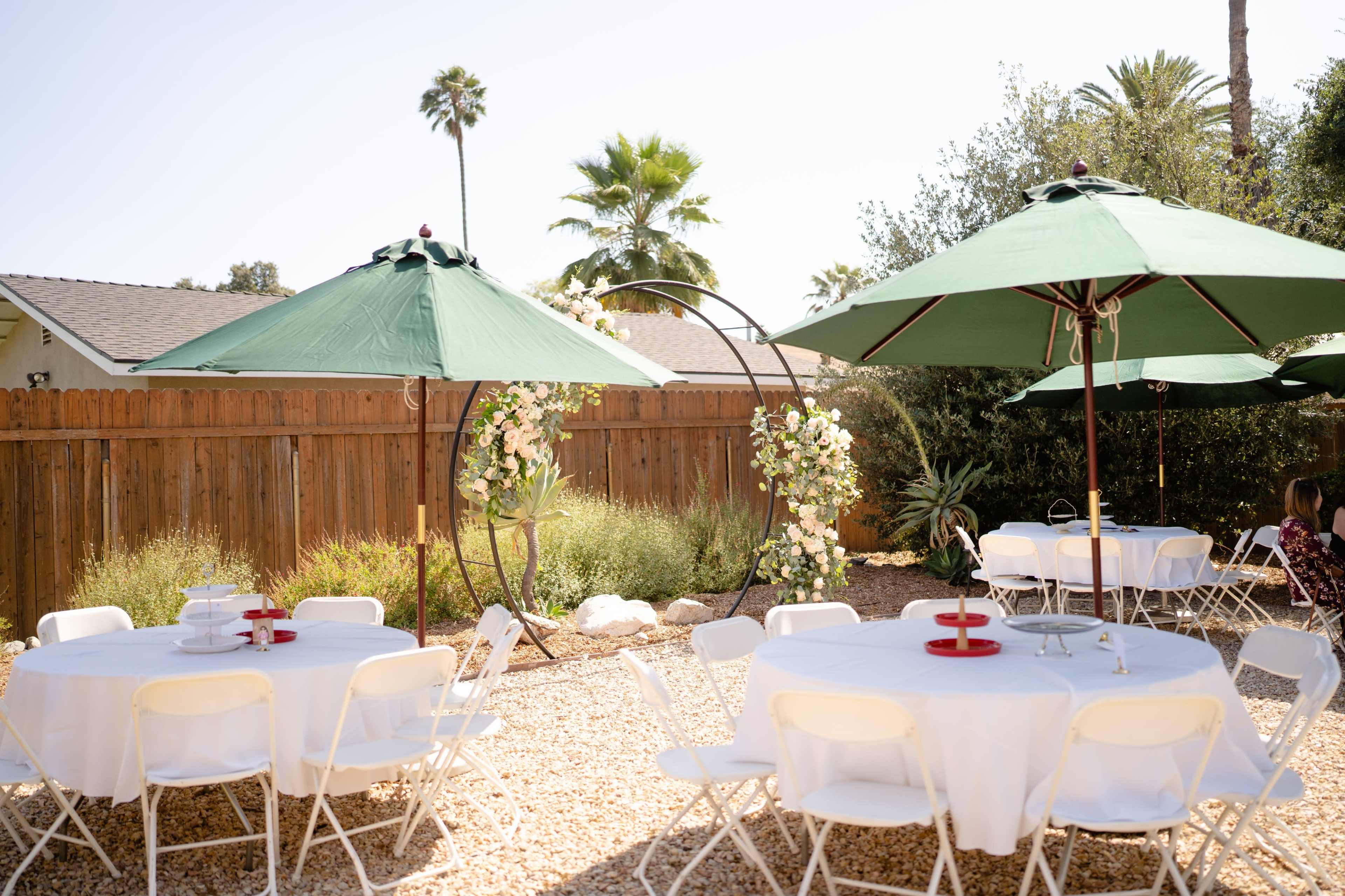 A backyard is set up for an outdoor gathering, featuring white tables and green umbrellas alongside greenery and a wooden fence.