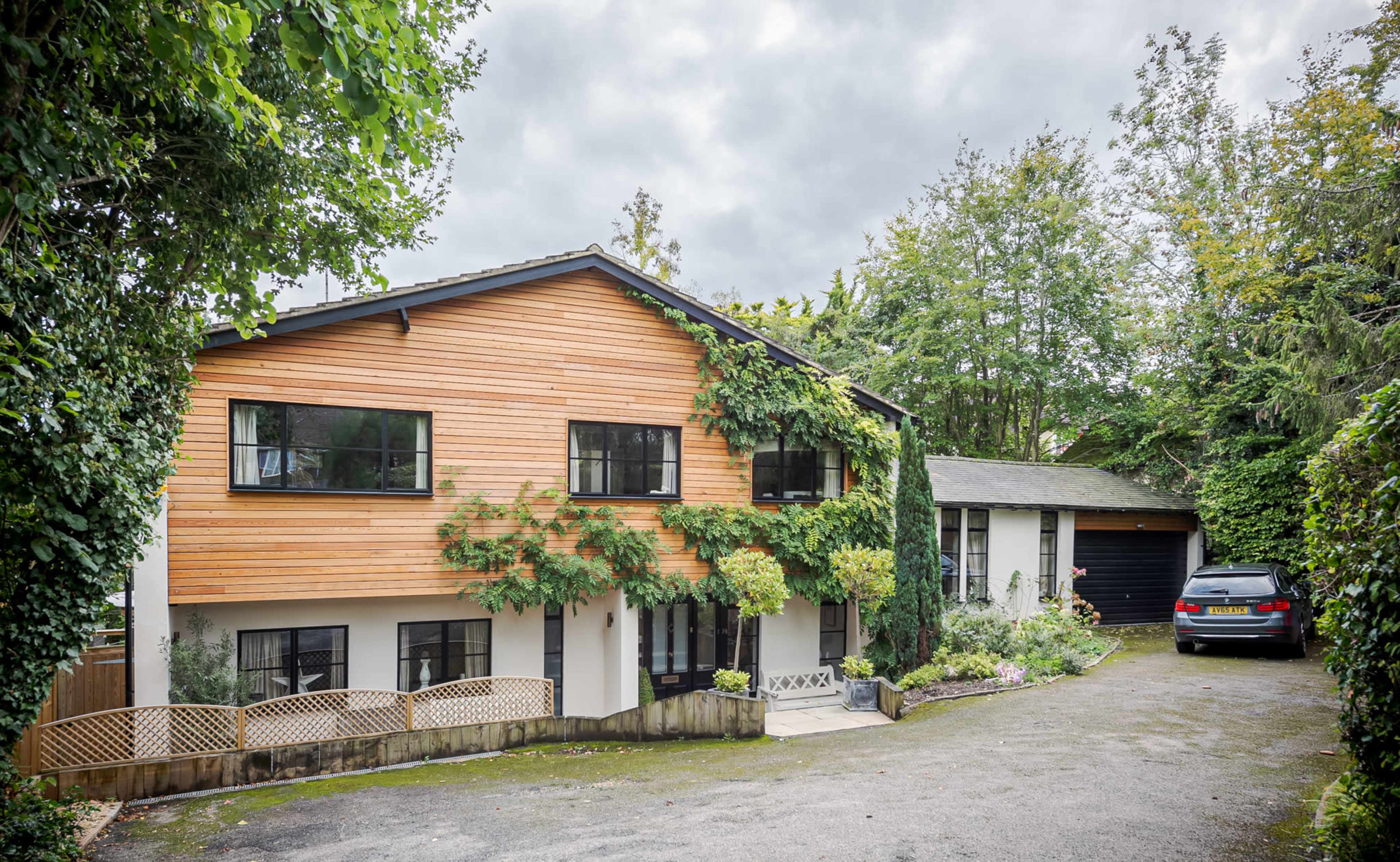 A two-story house with a wooden facade partially covered in greenery, surrounded by trees, with a driveway leading to a garage.