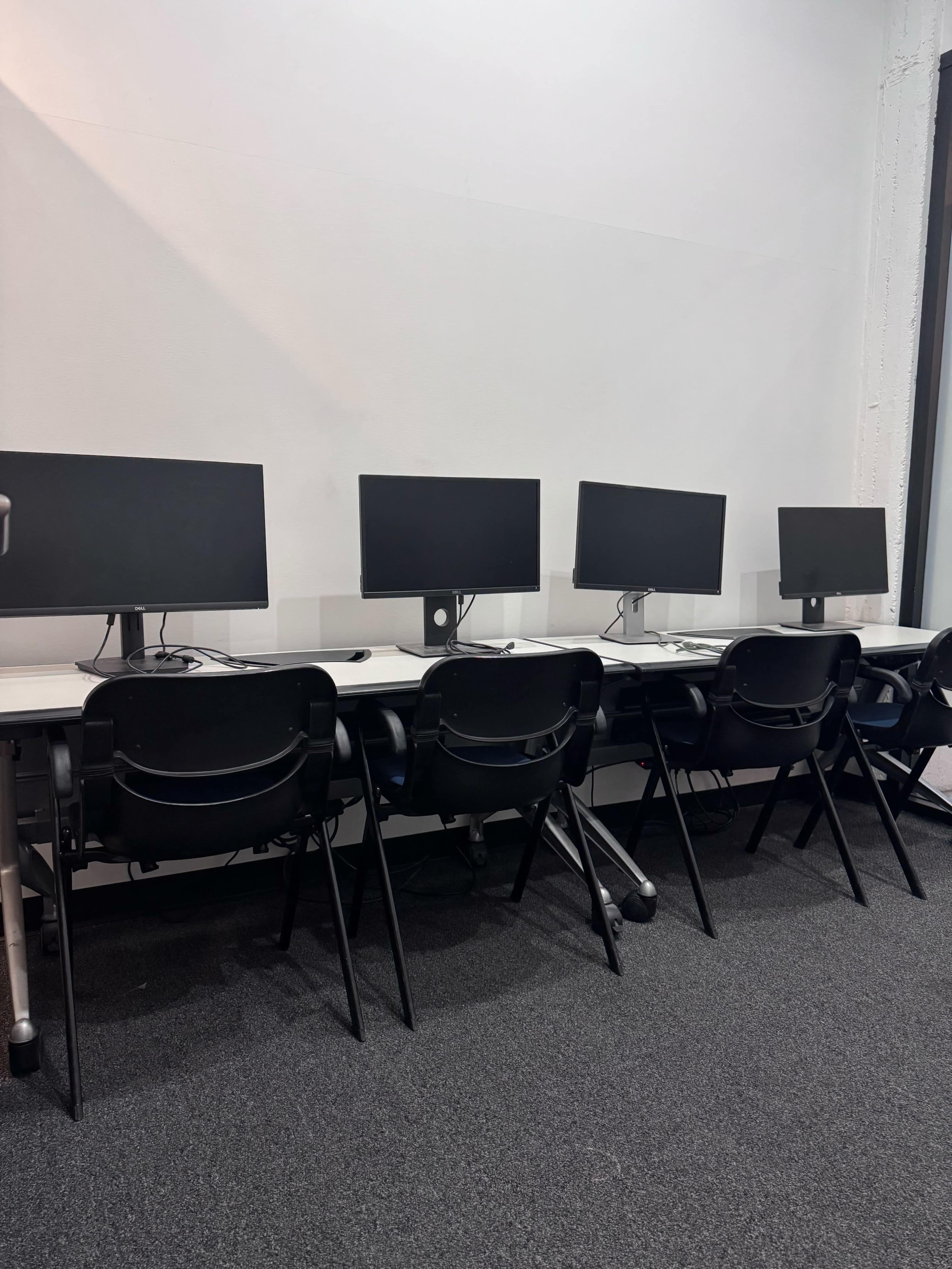 The image shows a row of four empty desks with computer monitors positioned on them in a modern office space.
