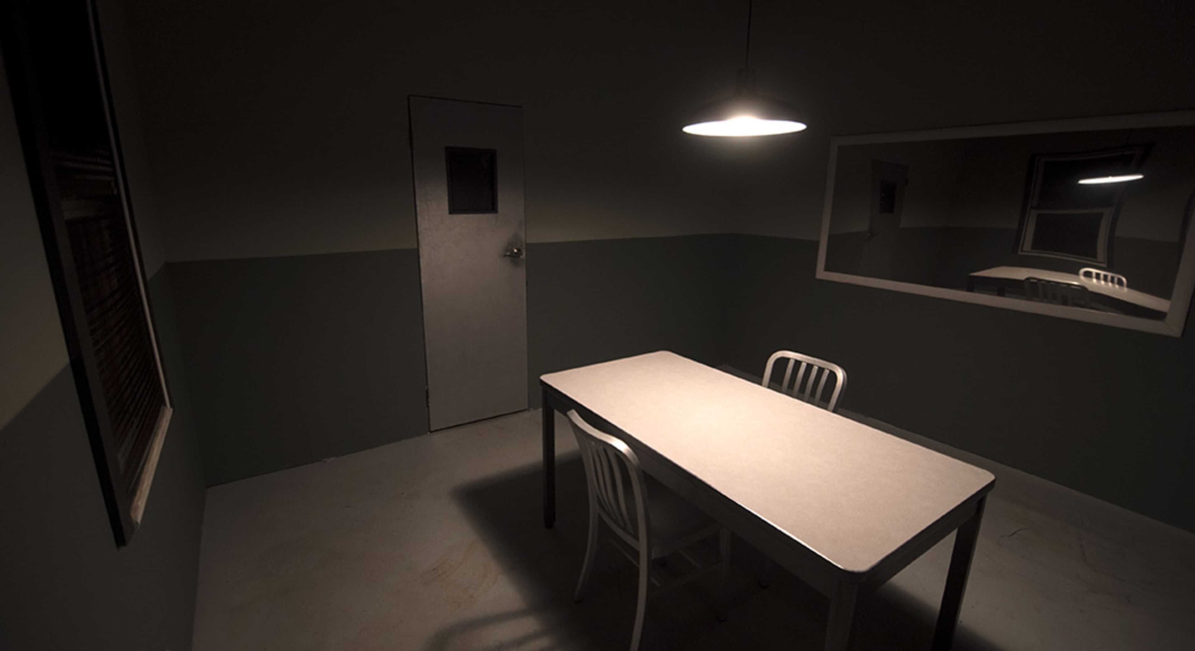 A dimly lit interrogation room features a table and chairs, with a door and a large mirror on the walls.