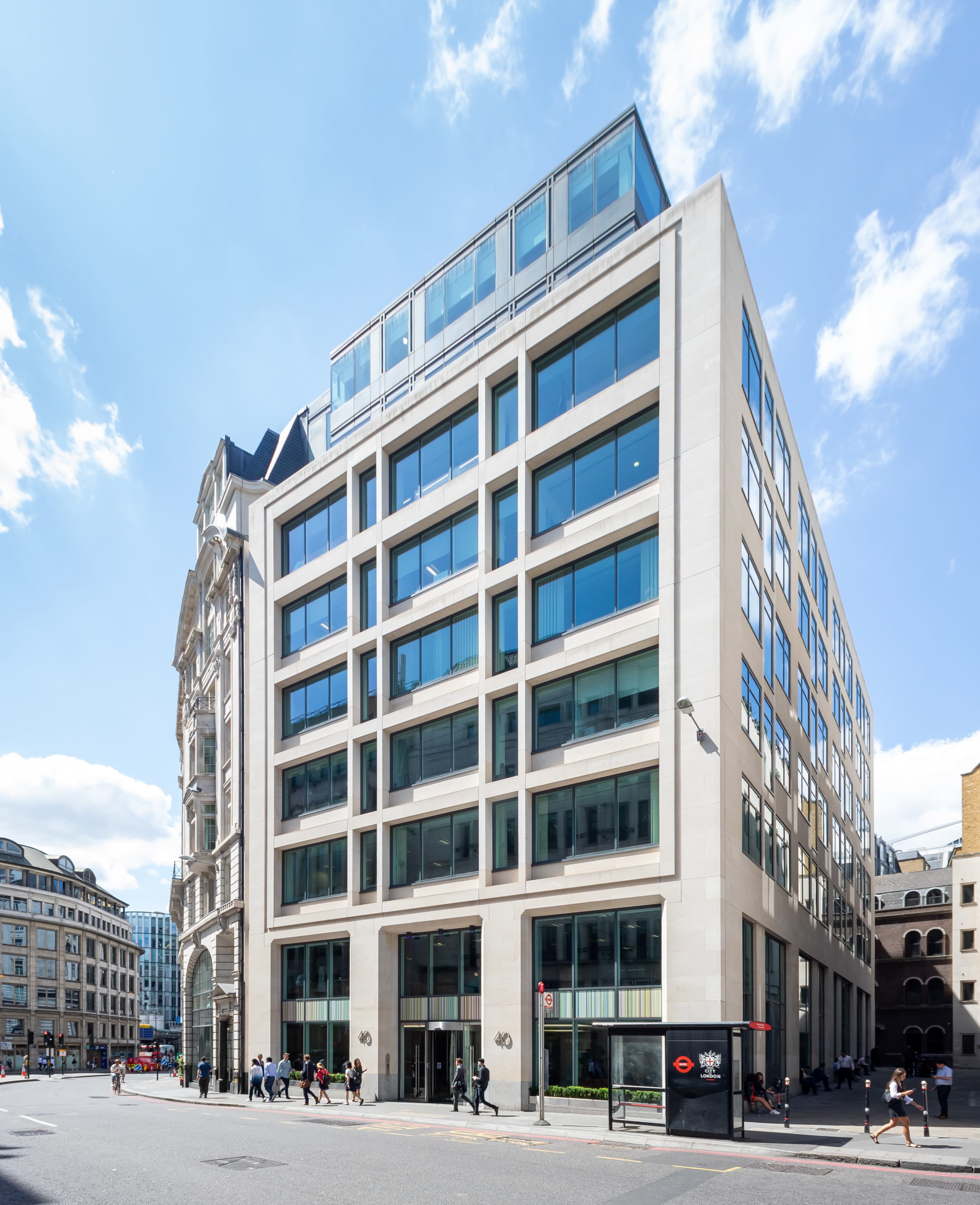 A modern multi-story building with large glass windows is situated at a street corner, with pedestrians walking by.