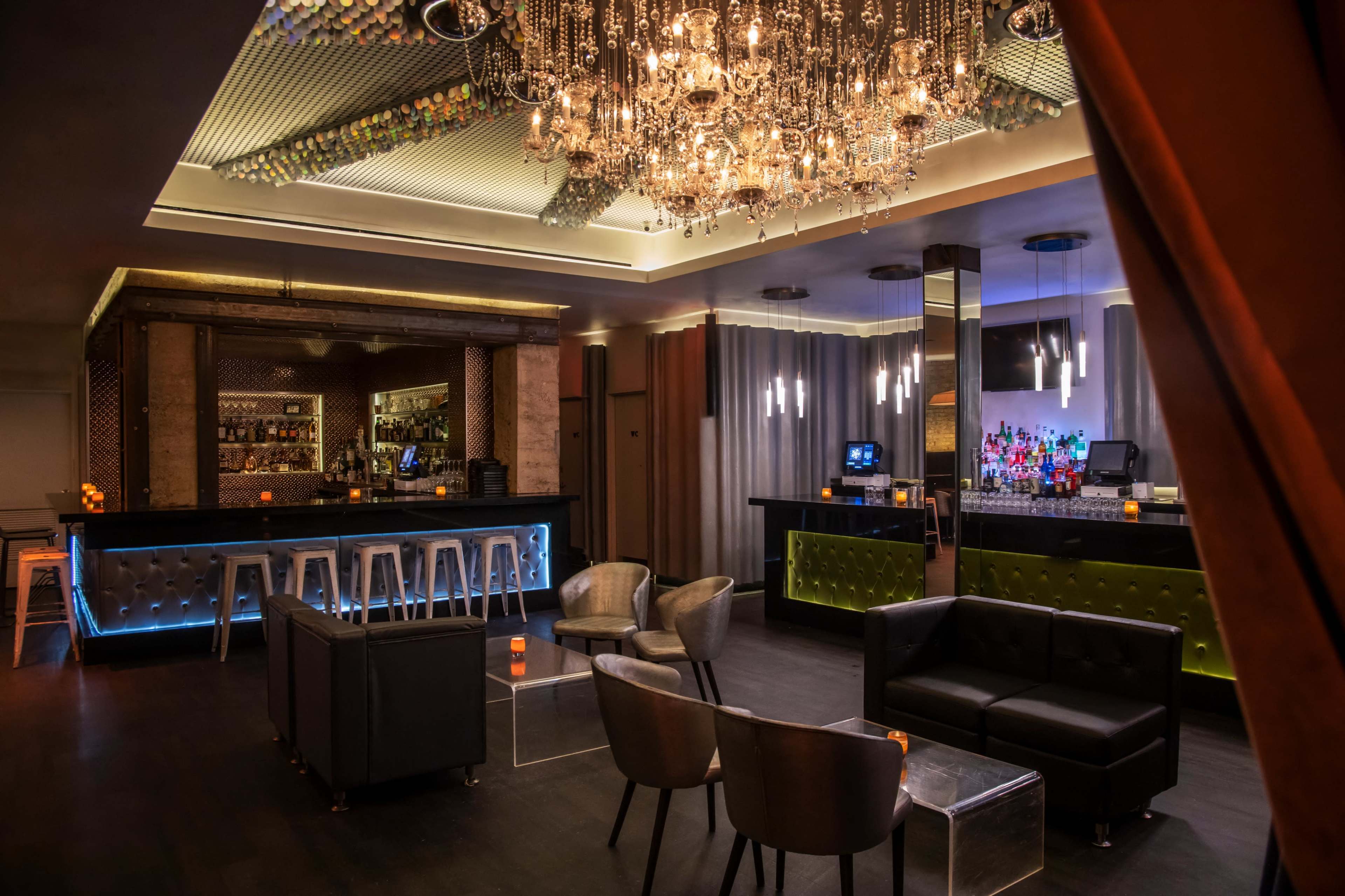 The image shows a modern bar area featuring a sleek black and chrome design with a chandelier overhead, illuminated shelves behind the bar, and various seating options arranged for patrons.