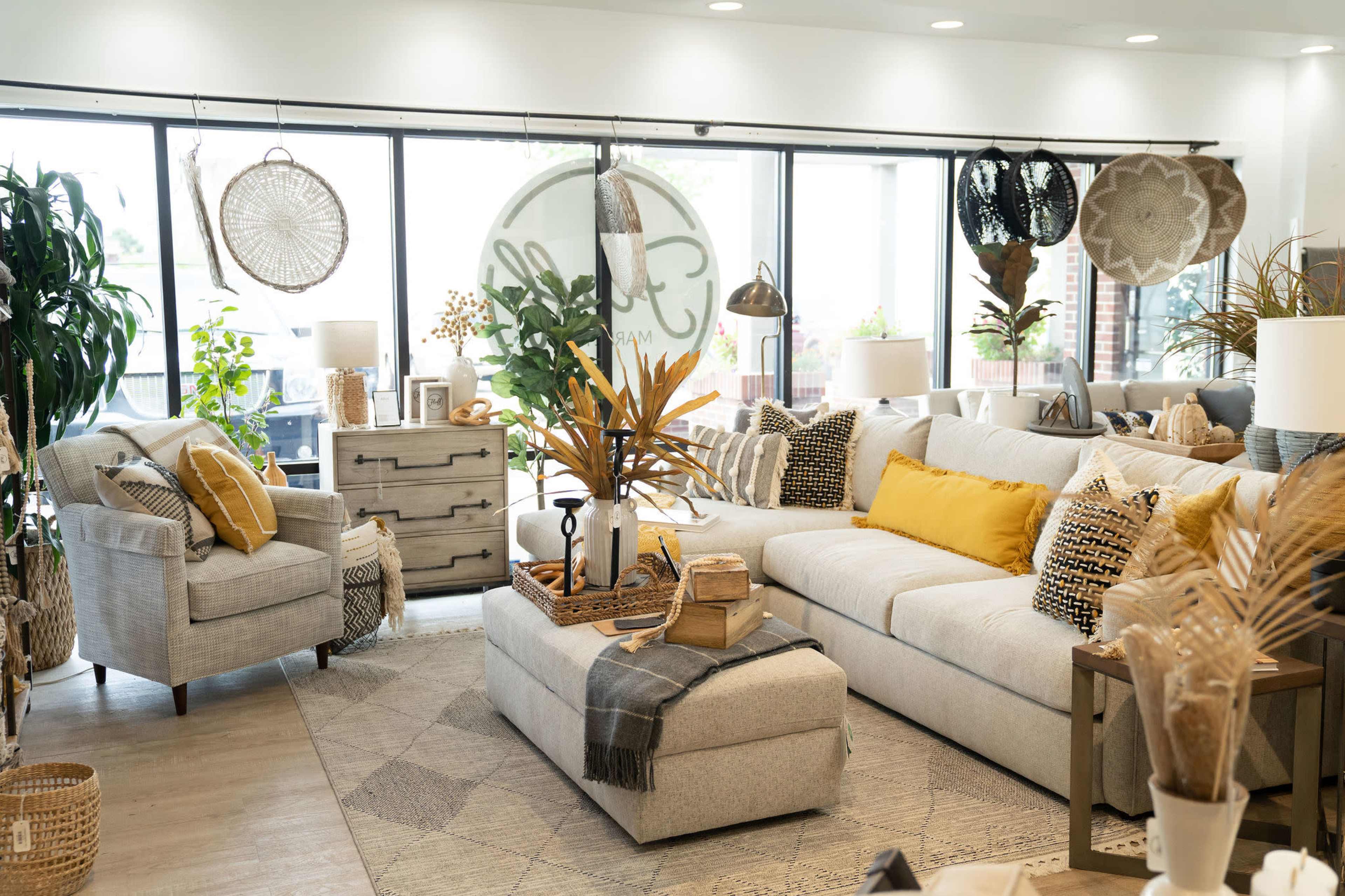 A modern living room features a spacious sectional couch with decorative pillows, a coffee table adorned with plants, and various textured furnishings beside large windows.