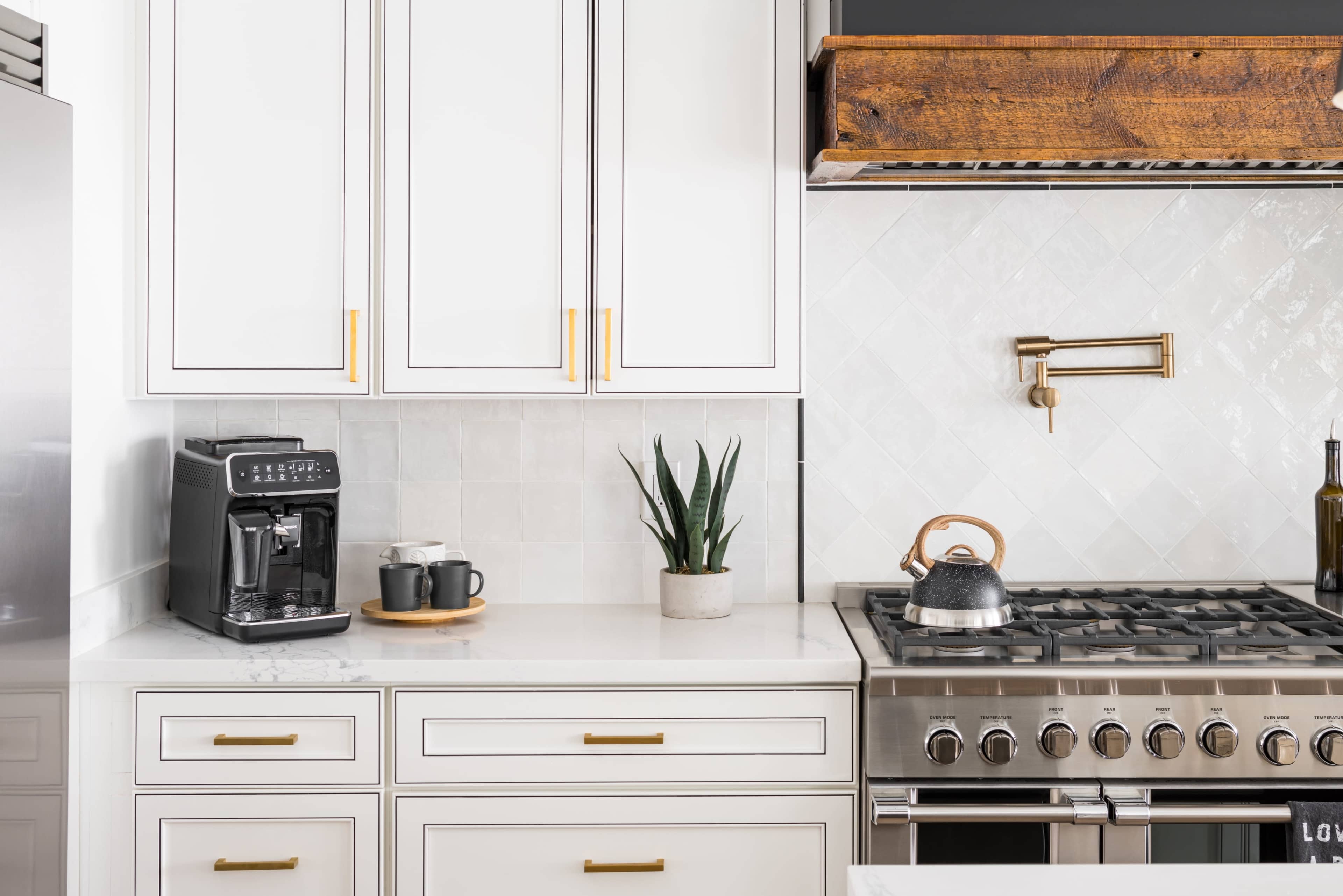 A modern kitchen features a countertop with an espresso machine, two mugs, a potted plant, and a gas stovetop with an overhead wooden vent.
