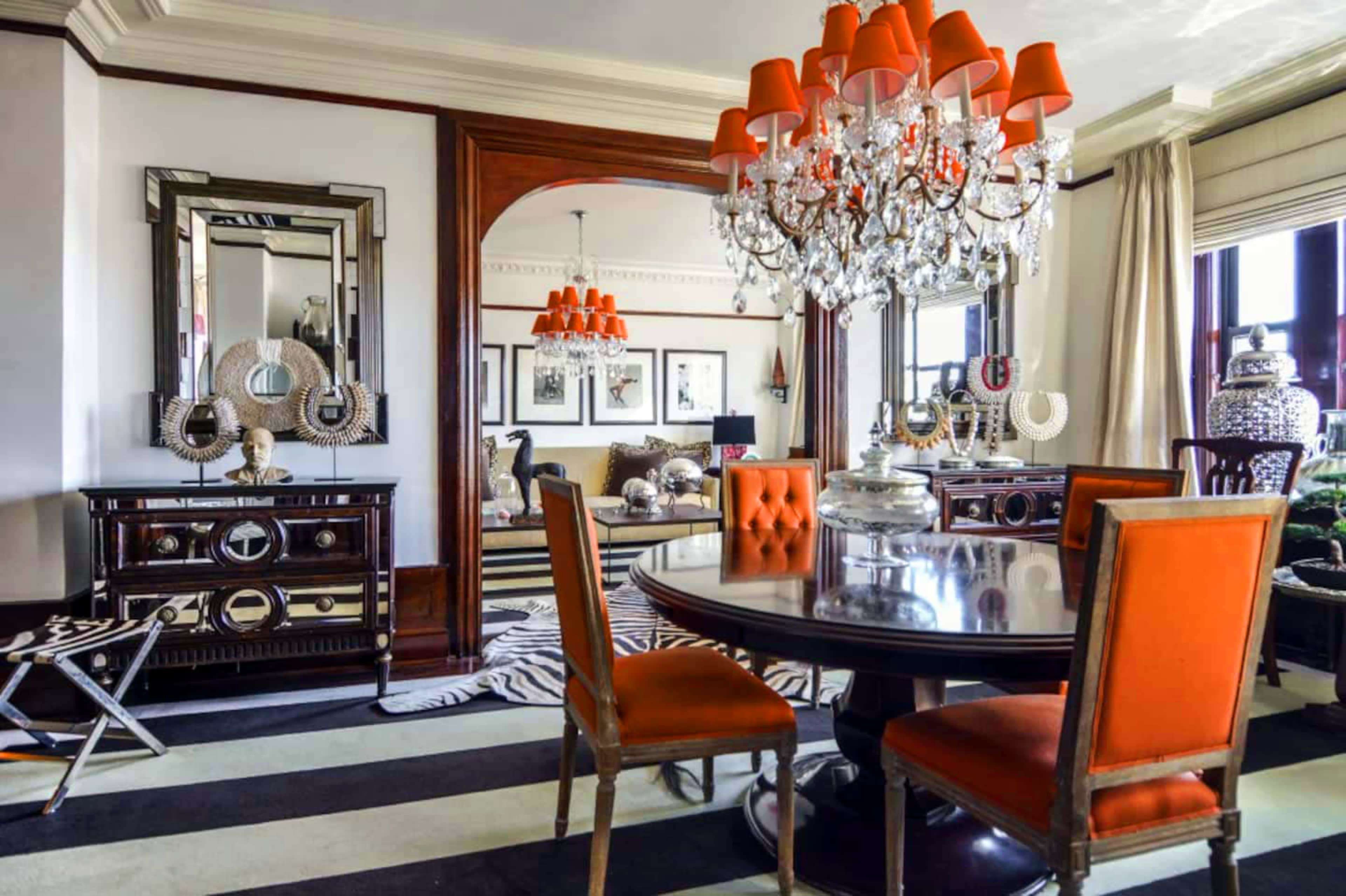 A stylish dining area featuring a round table with orange chairs, a crystal chandelier, and a black and white striped rug.