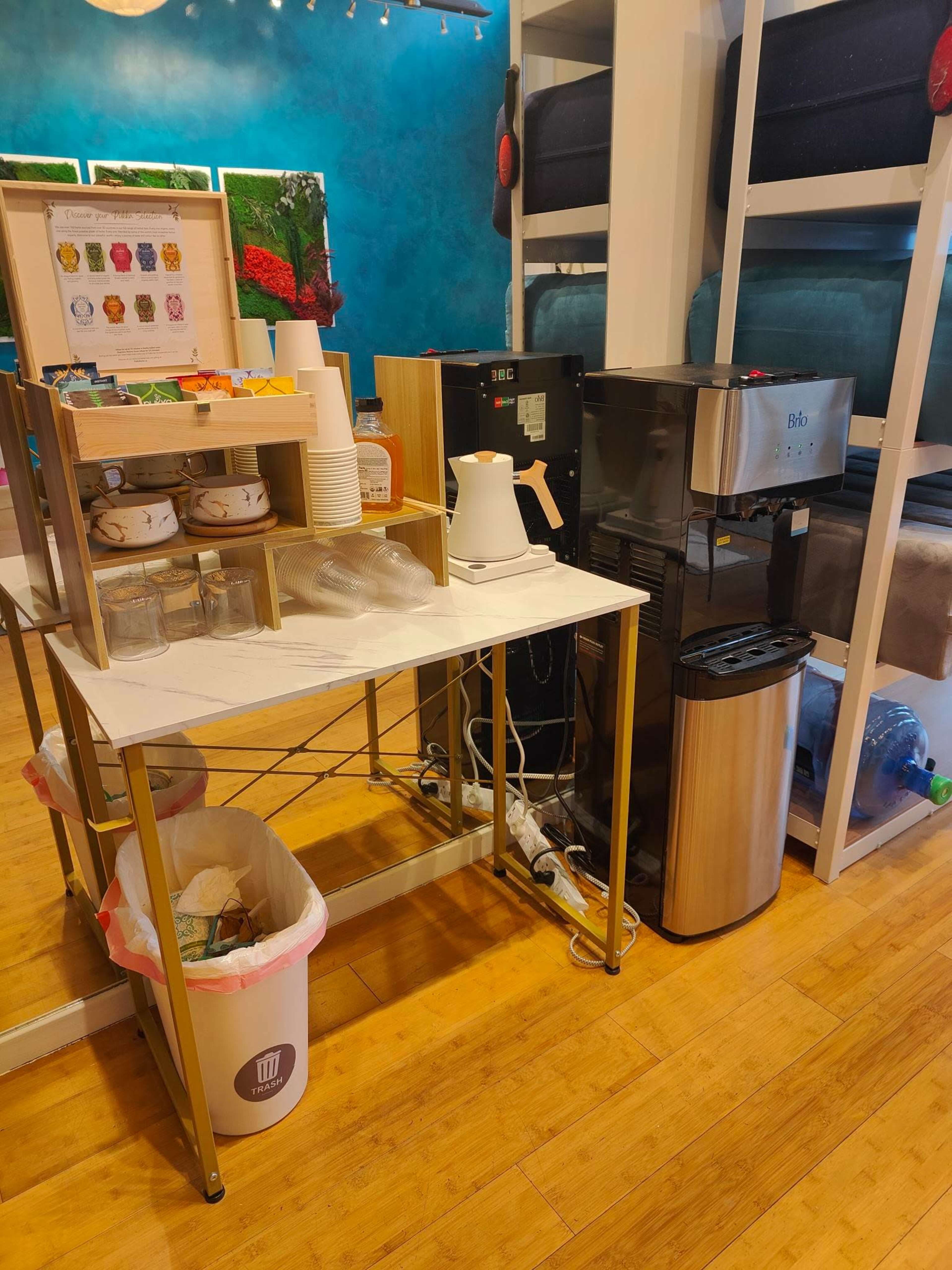A coffee station with a kettle, coffee machine, disposable cups, and a trash bin in a brightly colored room.