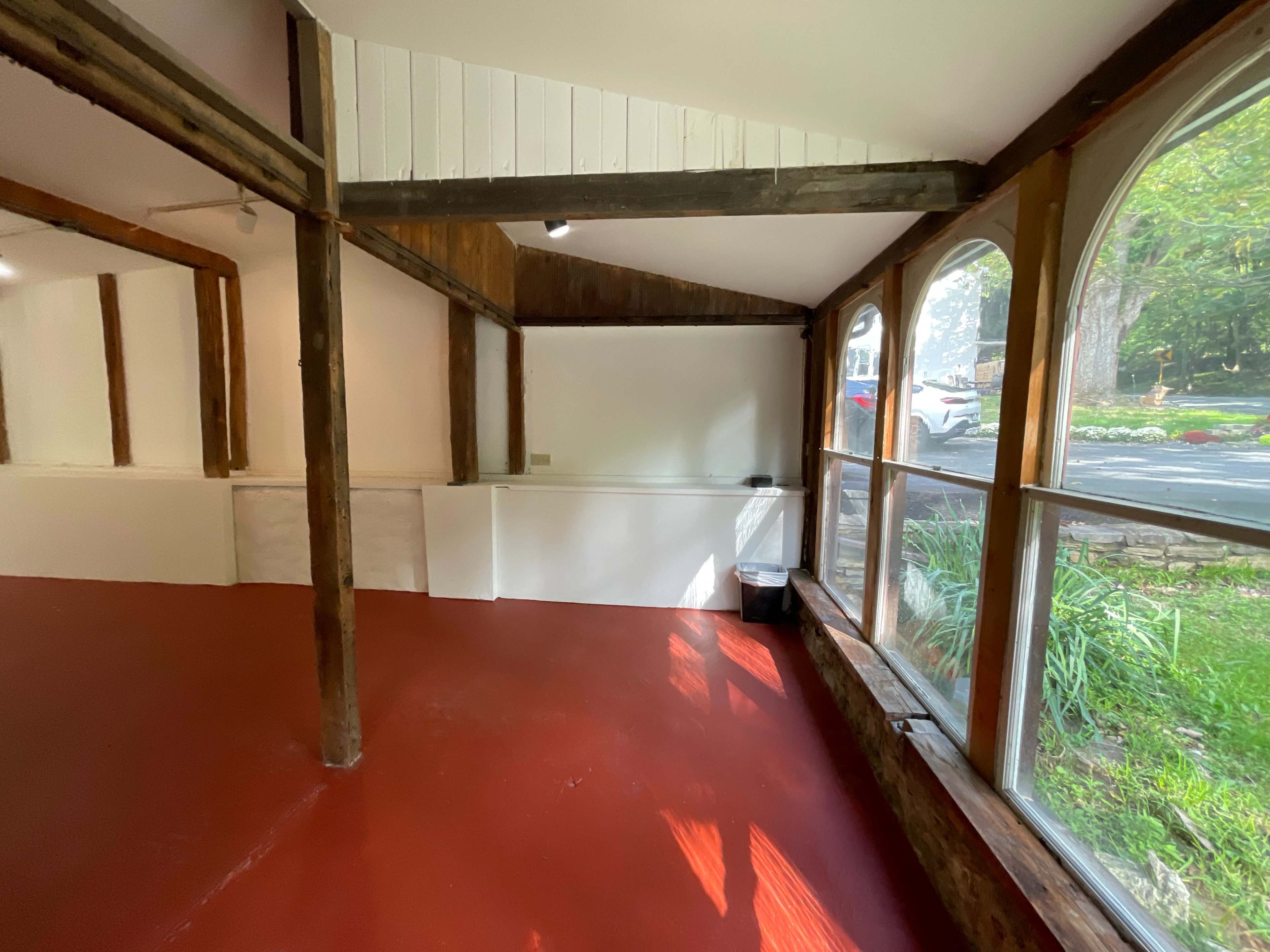 An interior space with wooden beams, a red floor, and large windows facing a landscaped area outside.