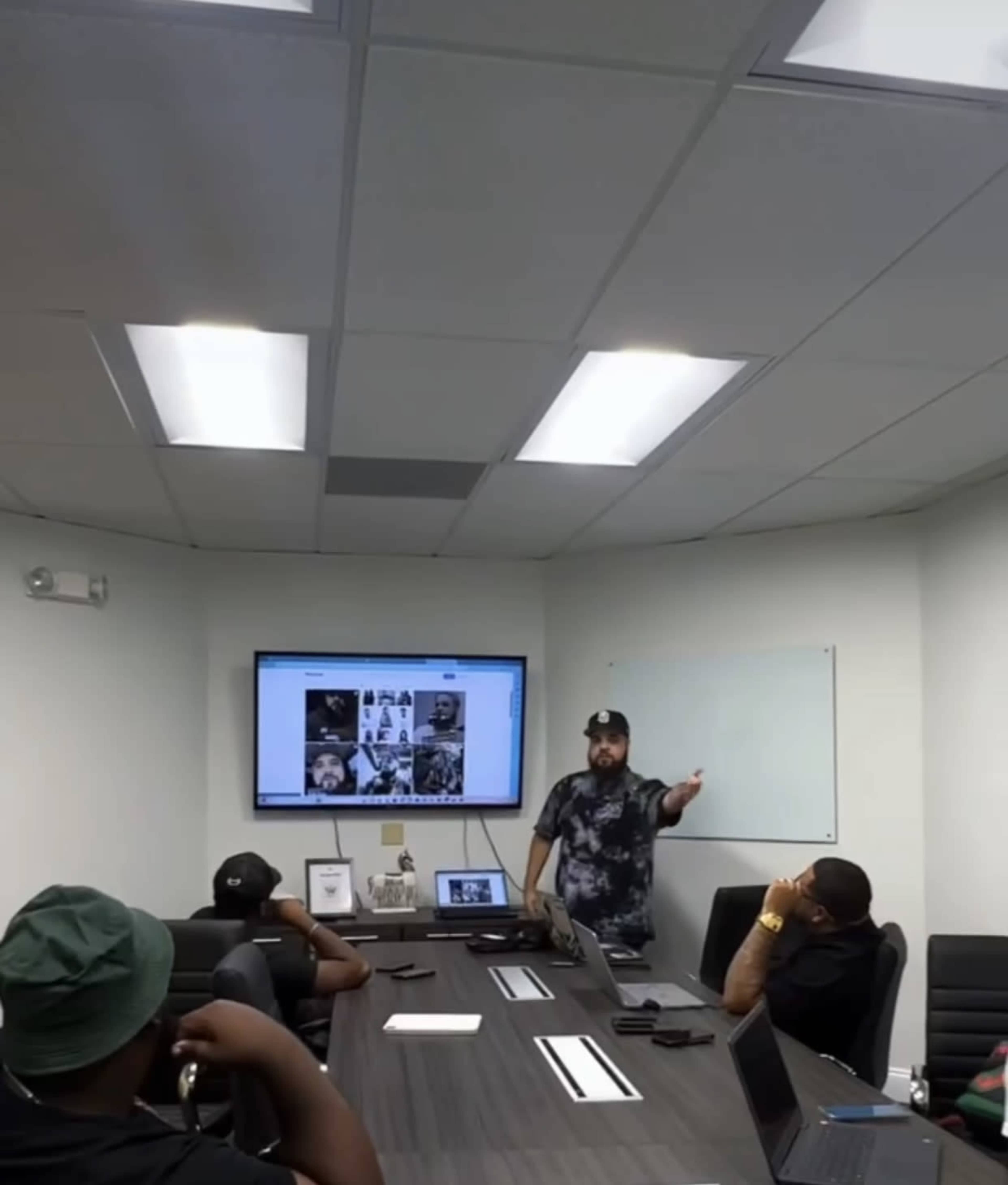 A man gestures while presenting to a group seated at a conference table in a meeting room.