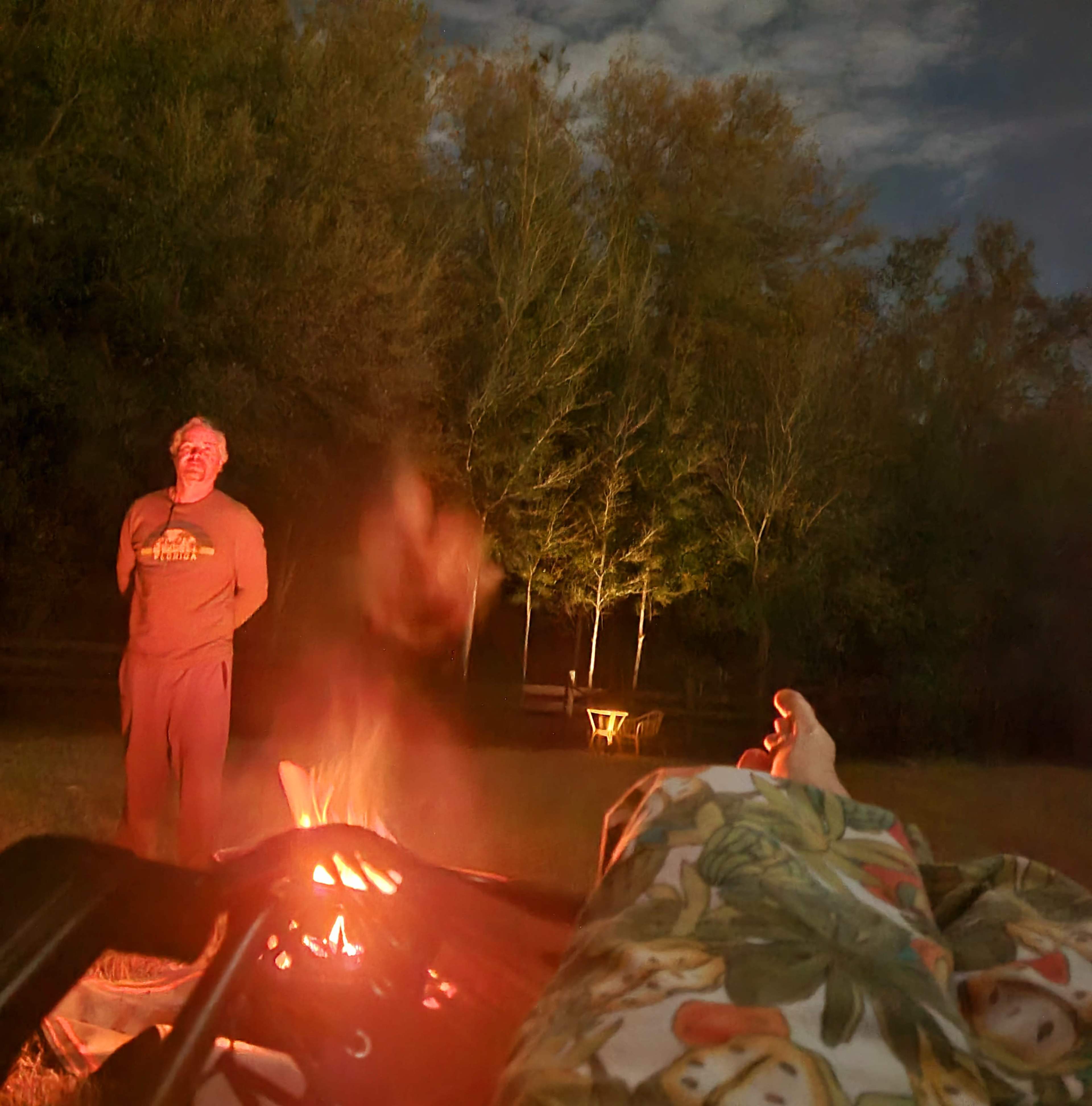 A campfire burns in the foreground with two people standing nearby under a night sky surrounded by trees.