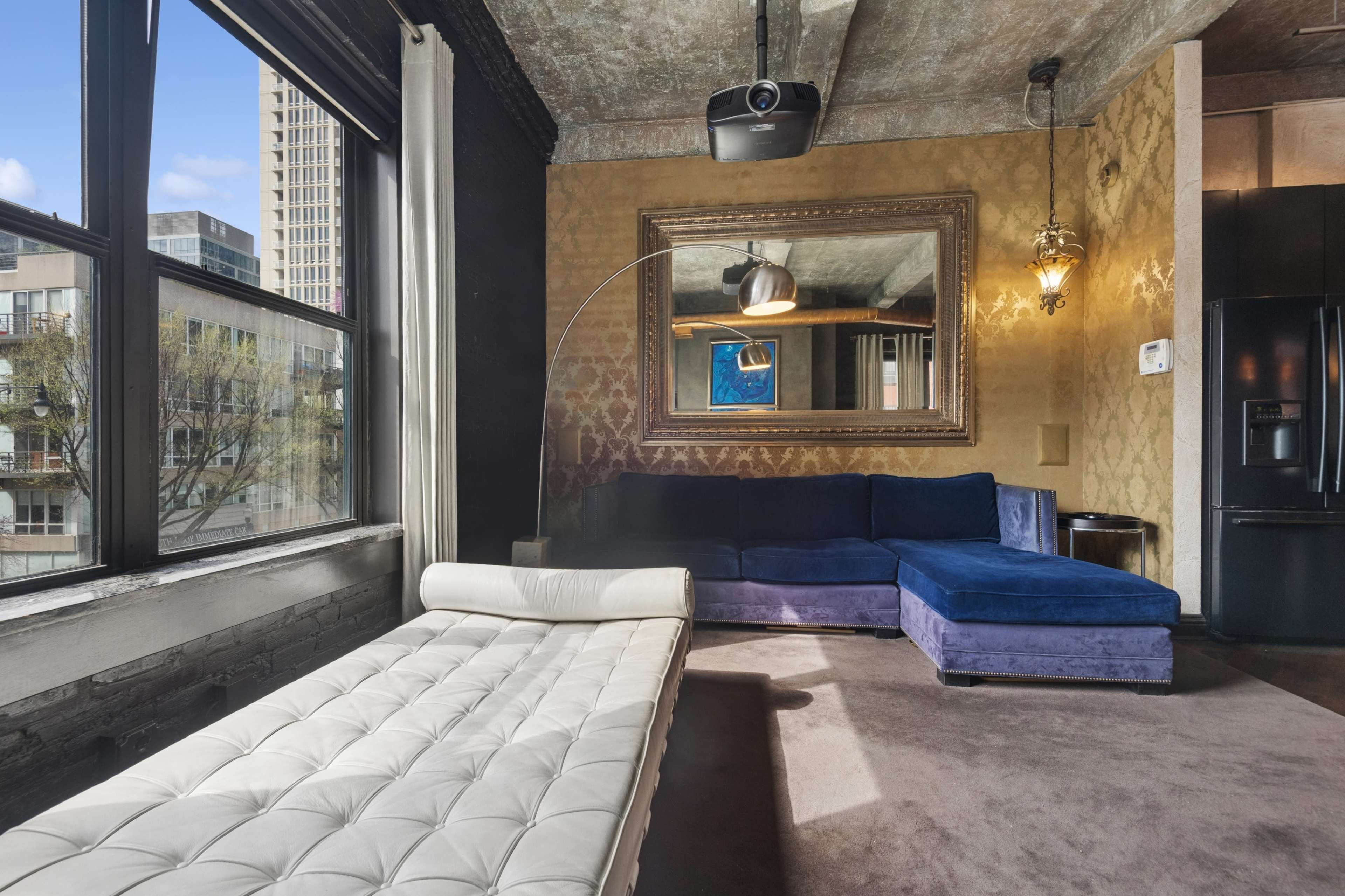 The image shows a modern living space featuring a white tufted lounge chair, a dark blue sectional sofa, large windows with city views, and a wall-mounted projector.