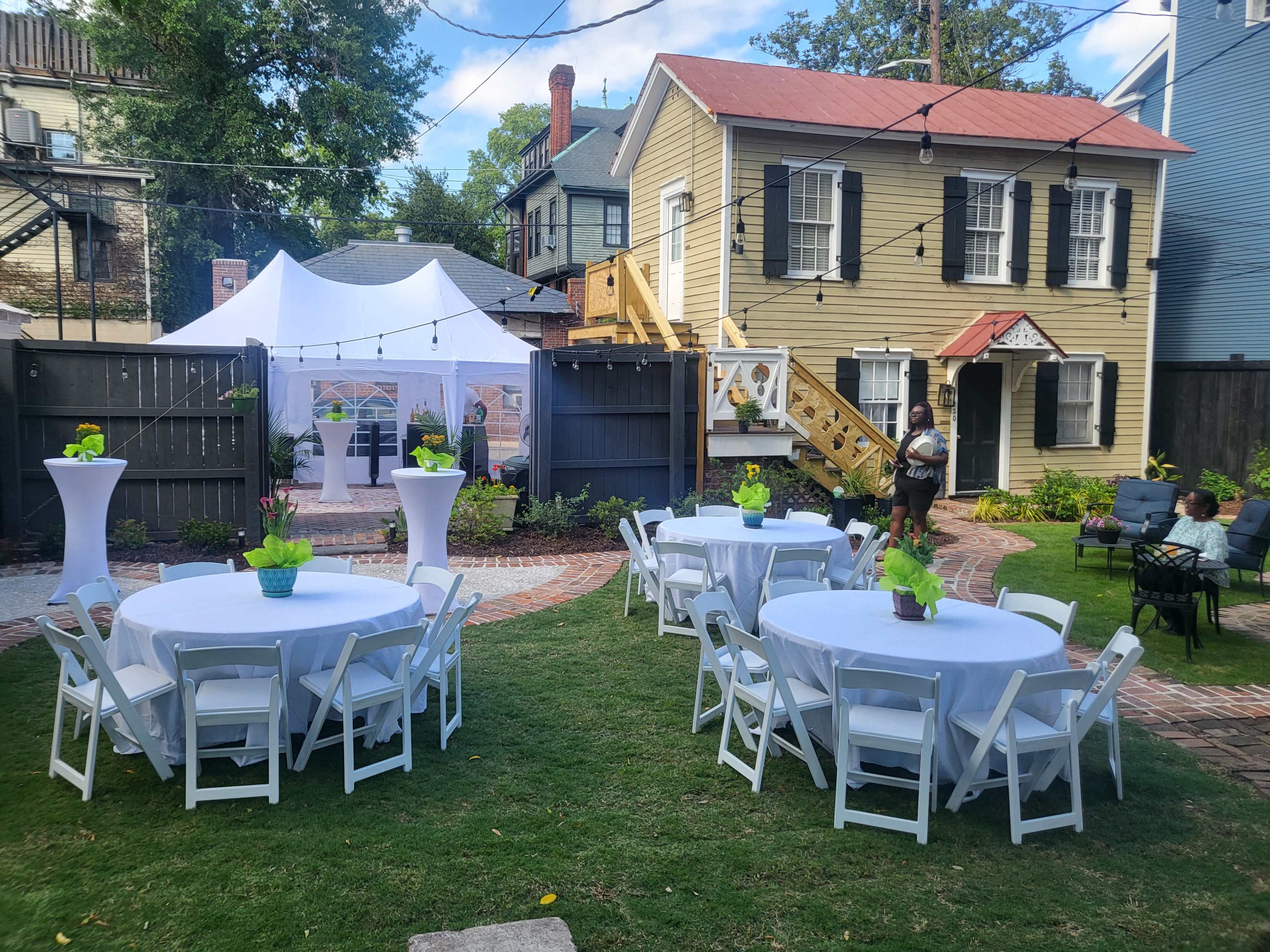 A courtyard setup for an event features round tables with white tablecloths and green centerpieces, surrounded by a white tent and a two-story house in the background.