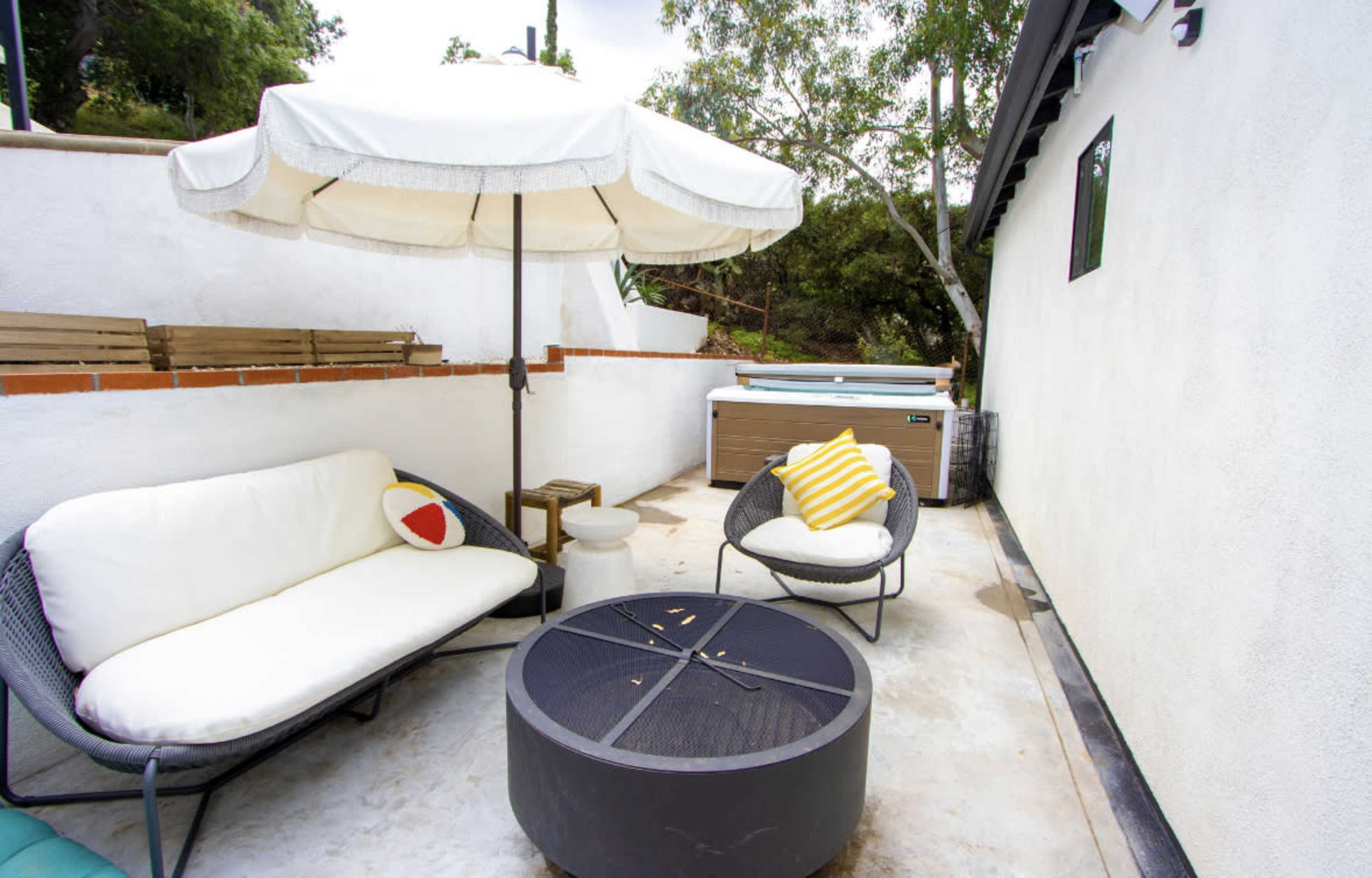 A small outdoor patio featuring a circular fire pit, a couch with pillows, a side table, and an umbrella for shade, alongside a hot tub against a white wall.
