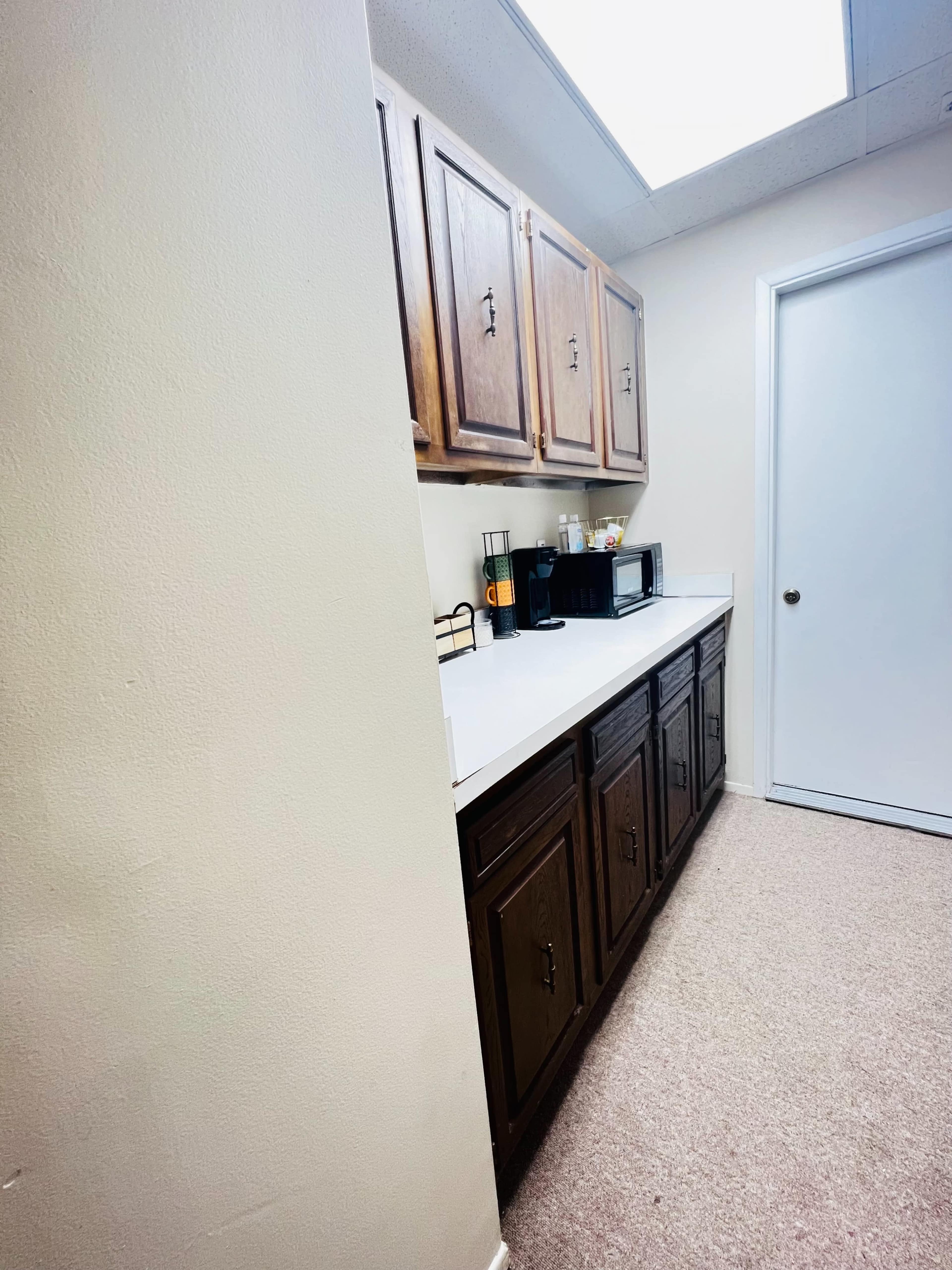 The image shows a narrow kitchen space with wooden cabinets above a white countertop and a microwave on the counter, leading to a closed door on the right.