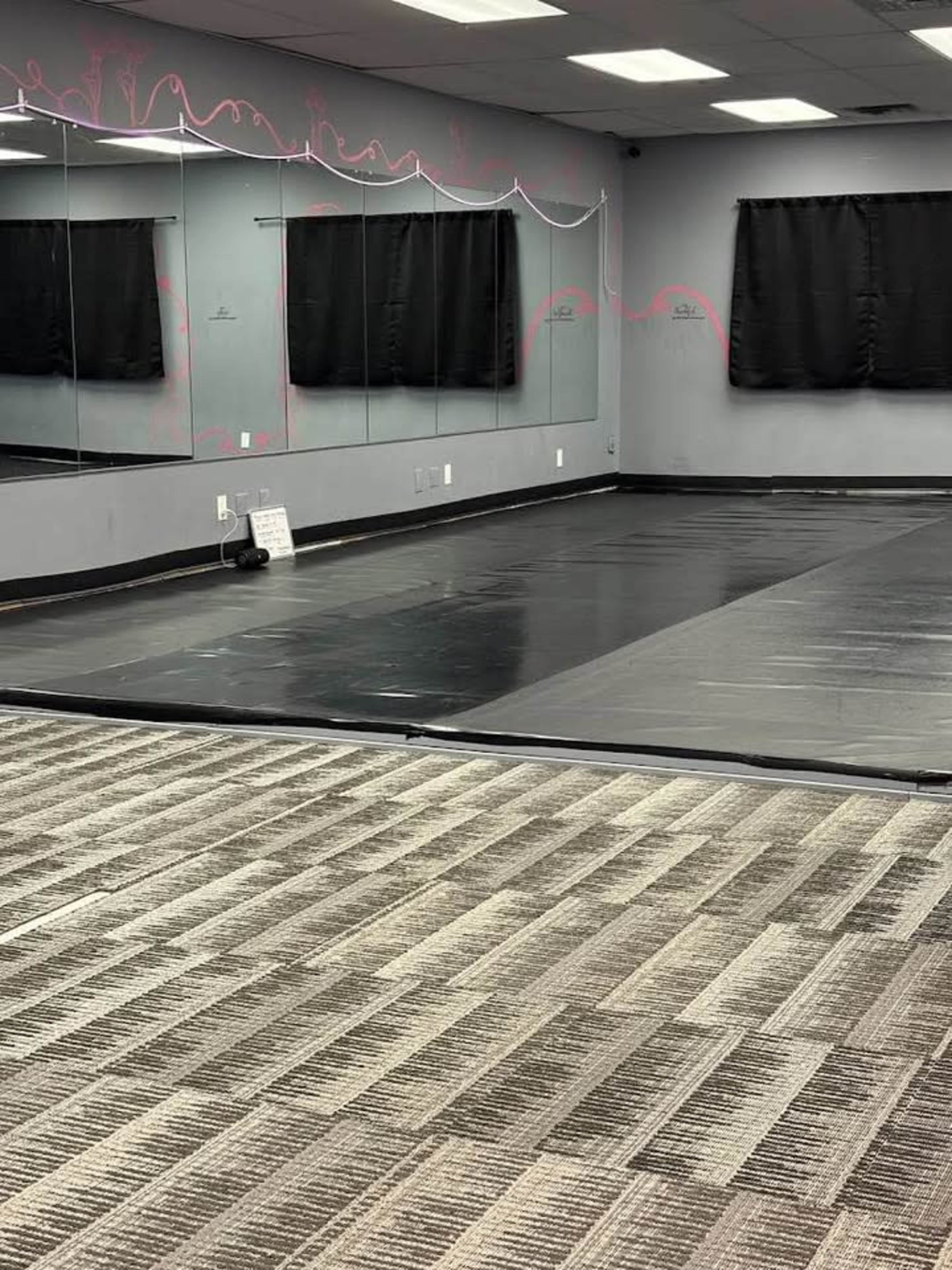 The image shows a dance studio with black flooring, mirrors on one wall, and windows covered by black curtains.