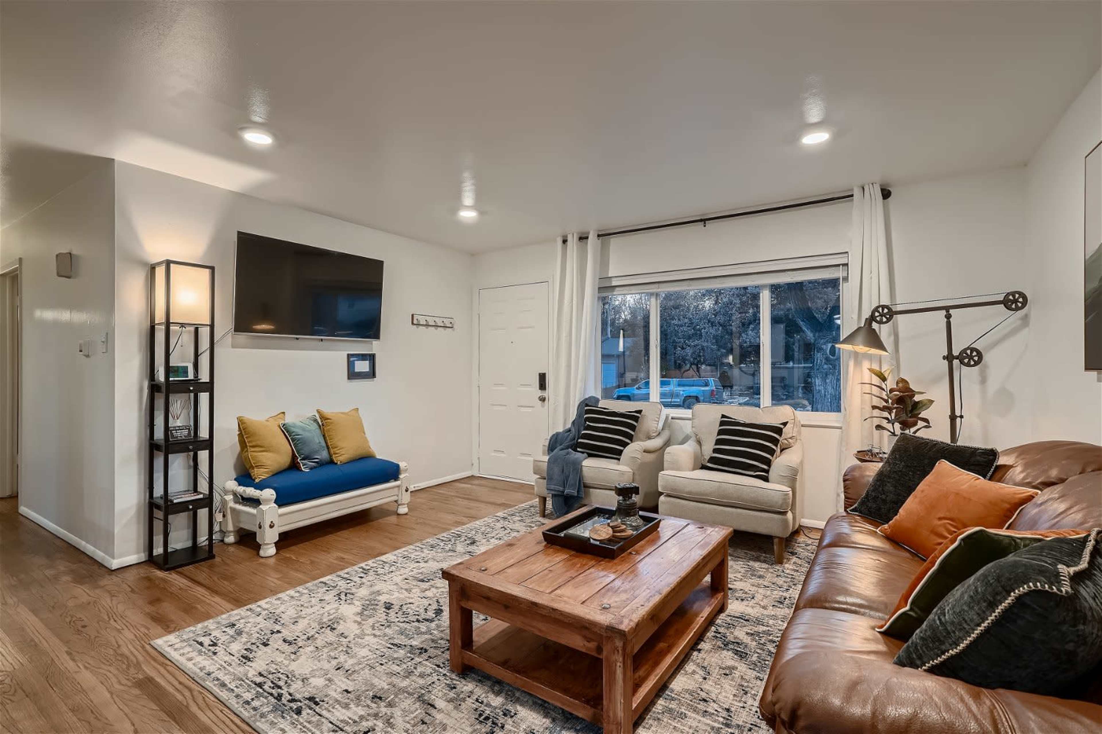 The image shows a modern living room with a brown leather sofa, a white armchair, a blue loveseat, a wooden coffee table, and a television mounted on the wall.