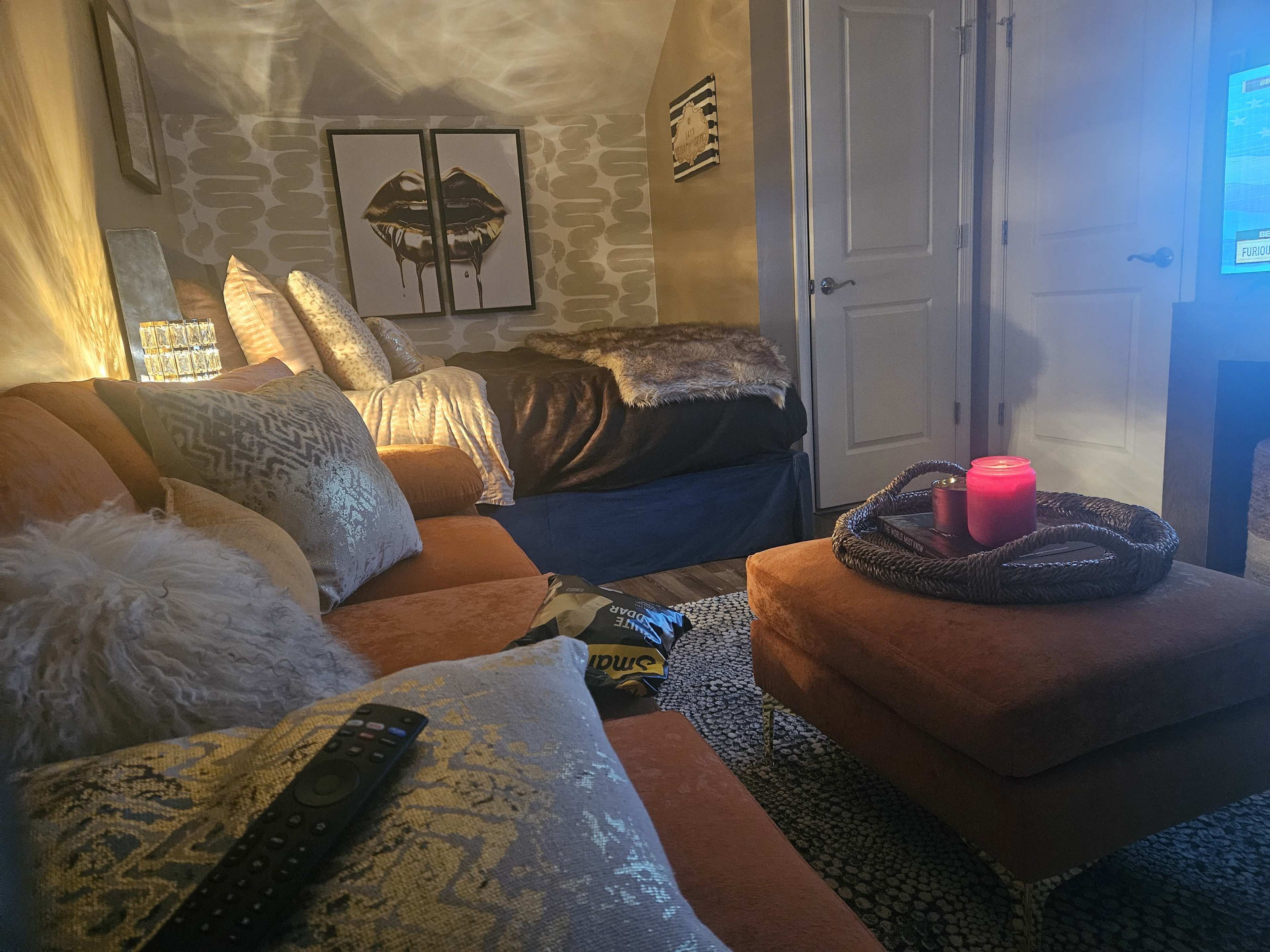 The image shows a cozy, softly lit room featuring an orange sectional sofa with decorative pillows, a small ottoman, and a bed in the background.