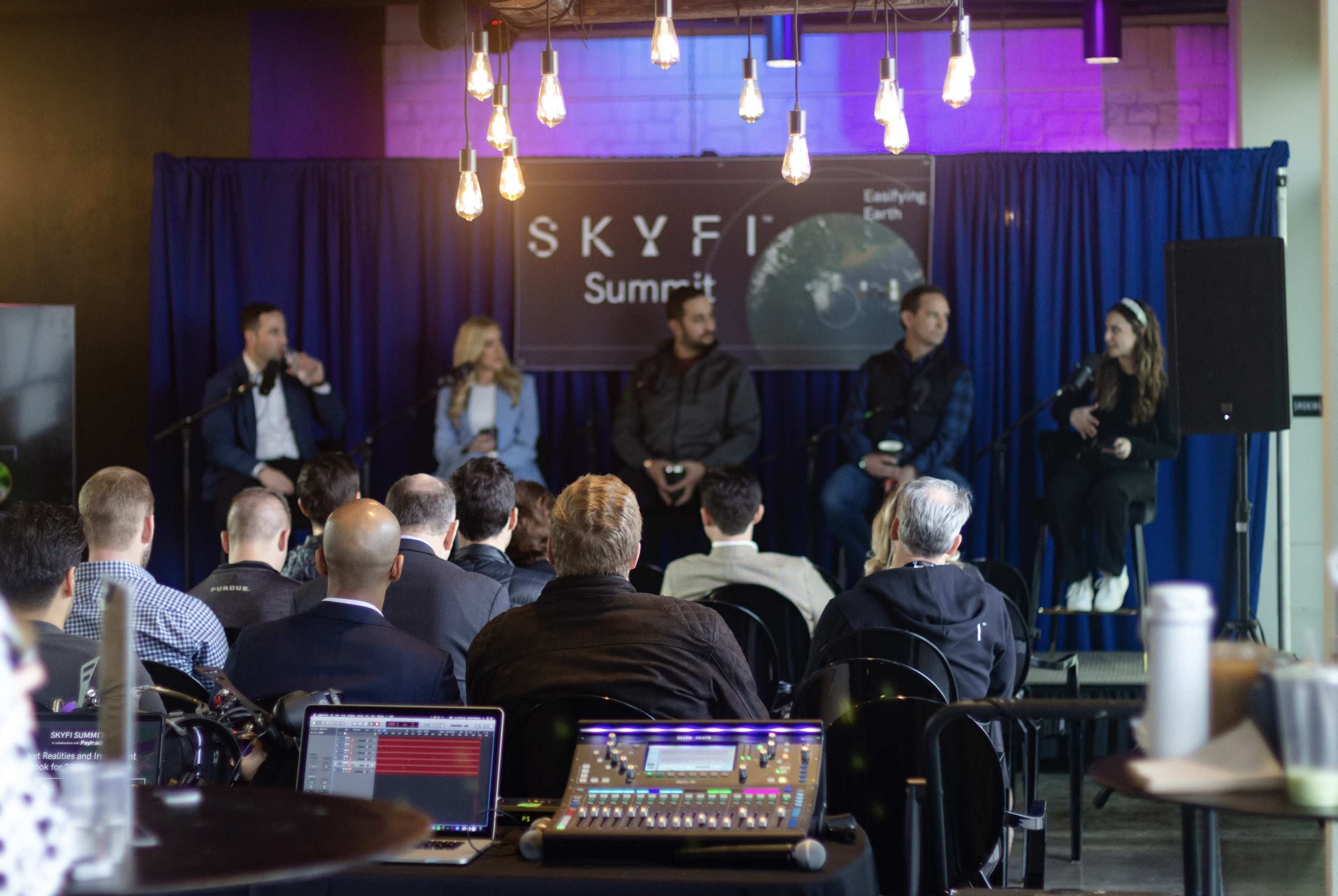 A panel discussion is taking place in a modern venue, with speakers seated on stage in front of an audience.