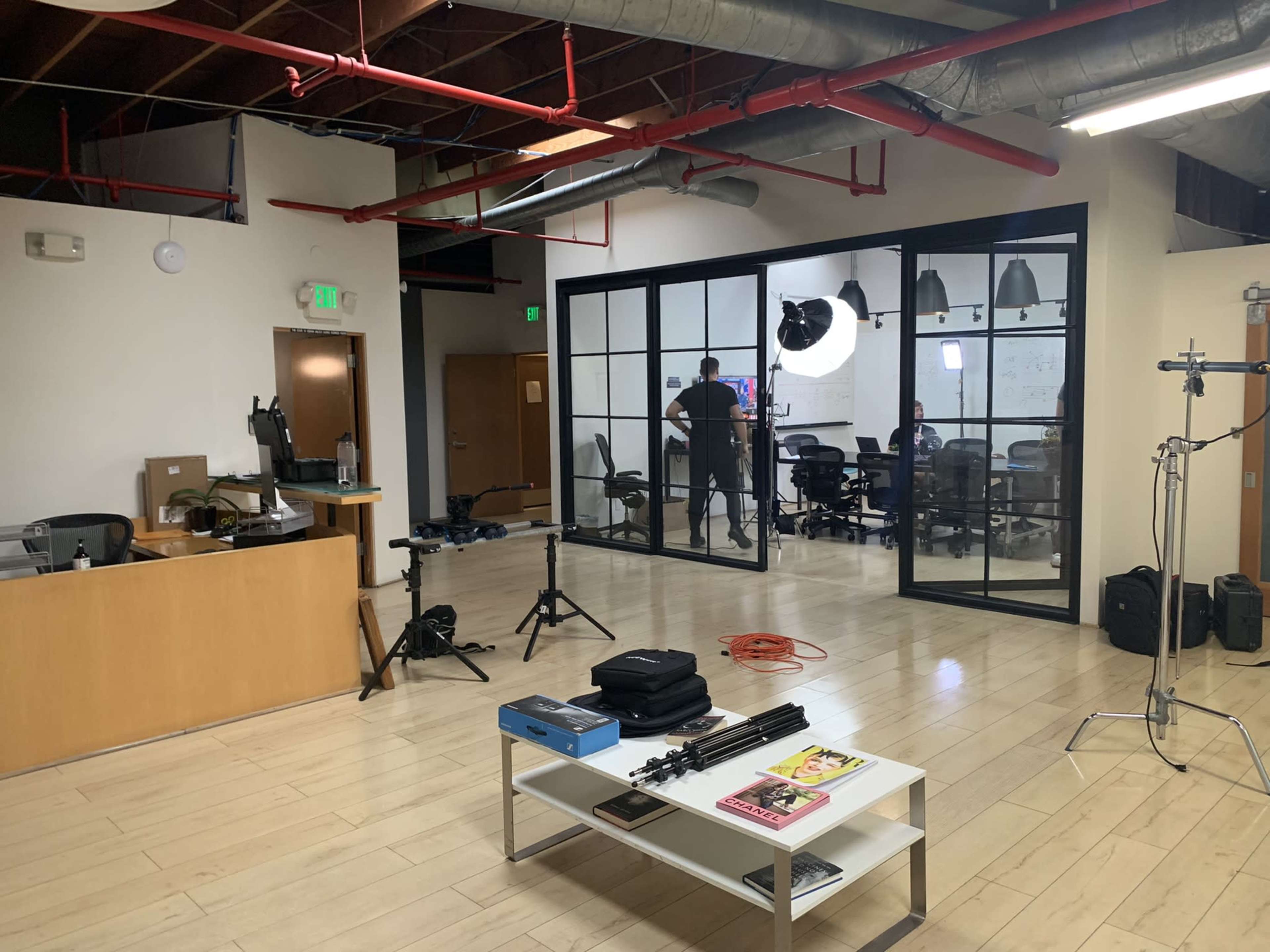 A modern office space features a reception area with a desk, a conference room in the background with people inside, and various equipment set up around the room.