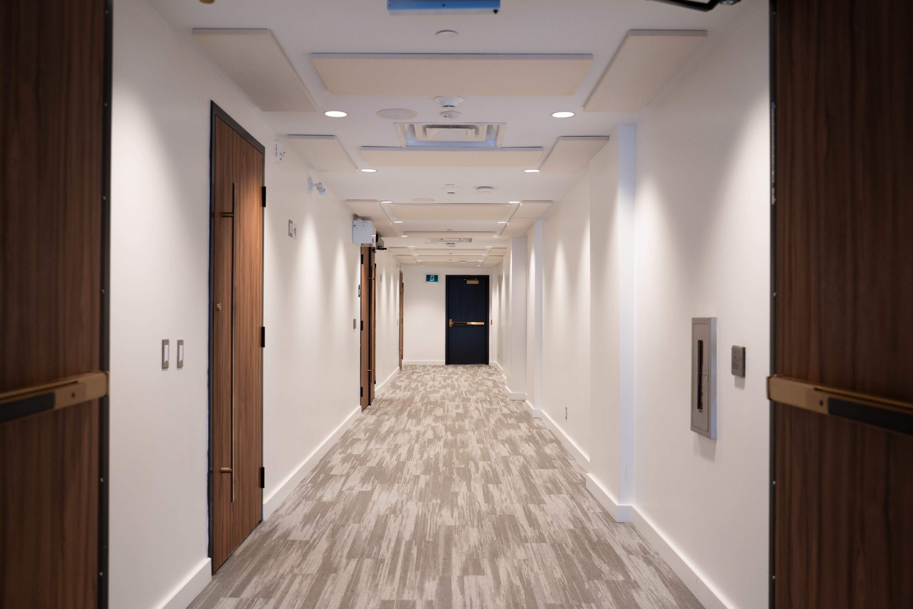 A well-lit, modern hallway features light-colored walls, a patterned carpet, and several doors lining each side.