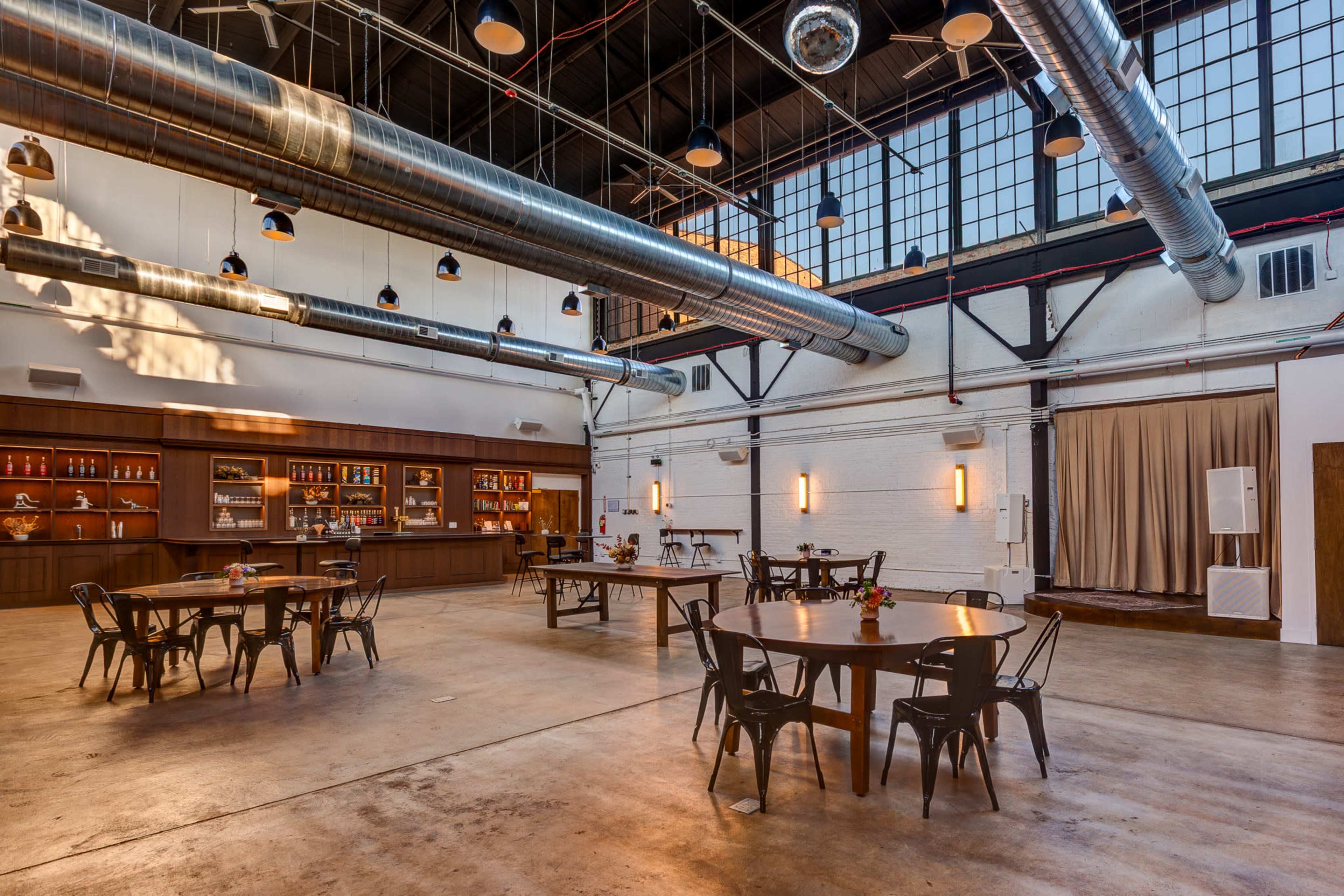 The image shows an industrial-style venue with wooden tables and black metal chairs arranged throughout the space, featuring large windows and exposed ductwork.