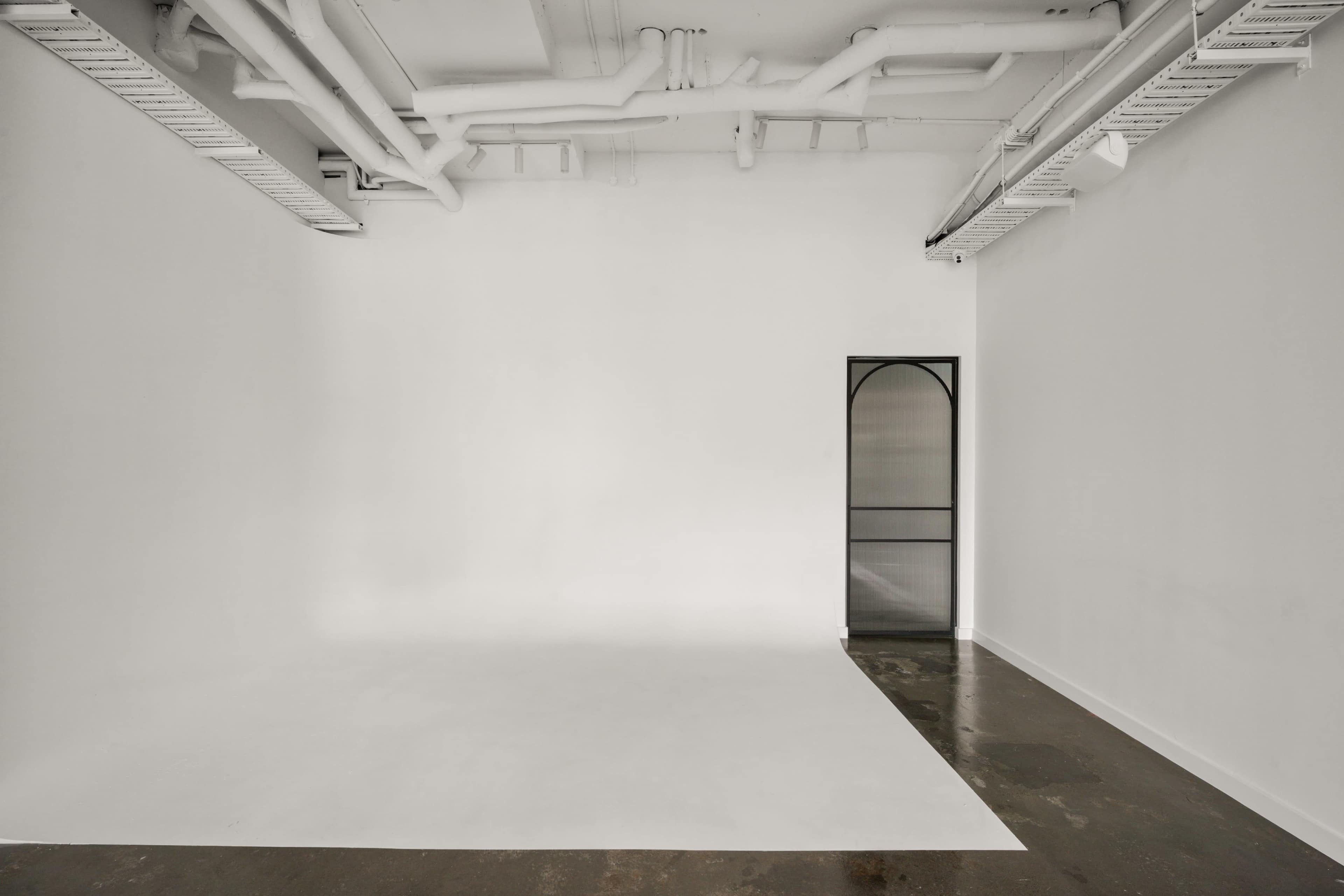 The image shows a minimalistic, empty room with white walls, a concrete floor, and a door featuring an arched design.