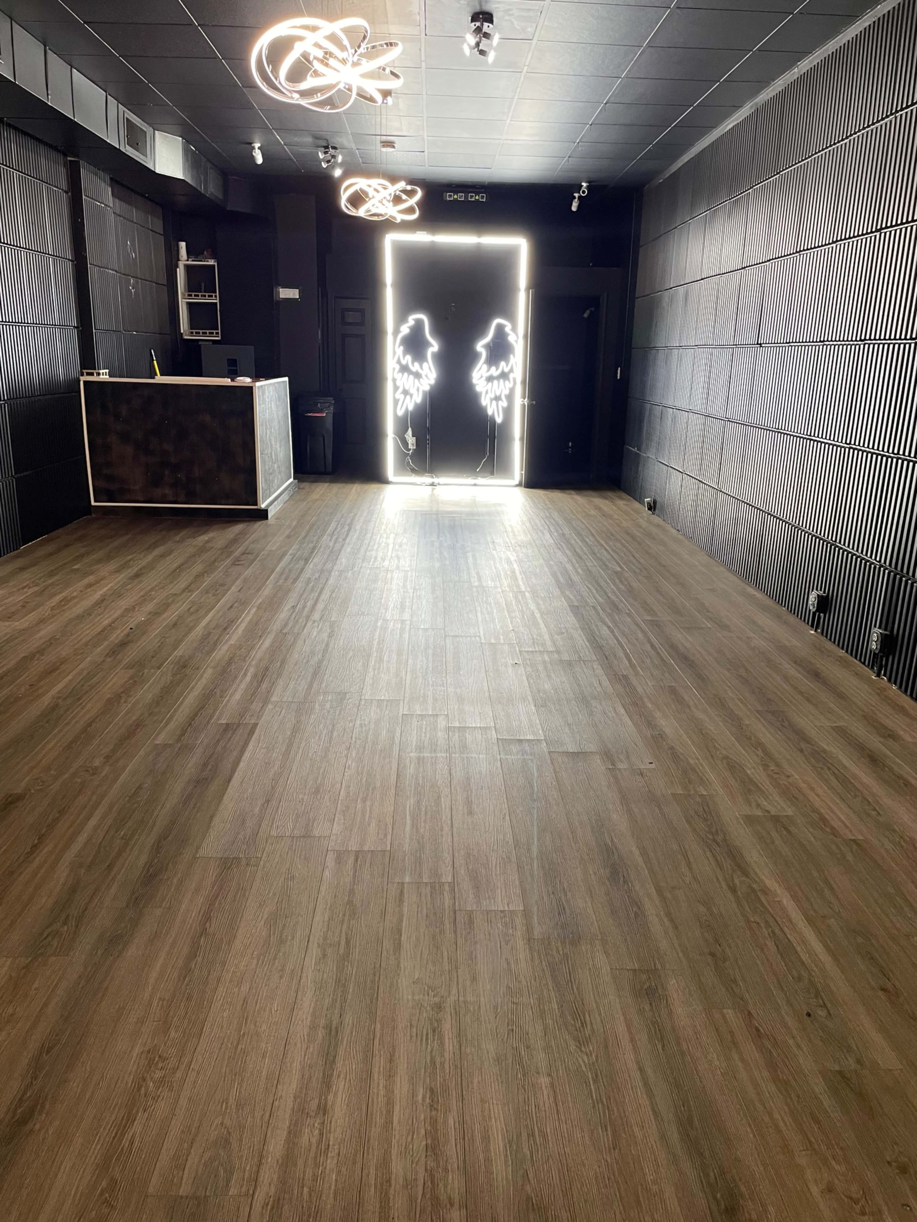 The image shows a spacious room with wooden flooring, featuring a reception area on the left and a brightly lit entrance with neon wing designs at the far end.