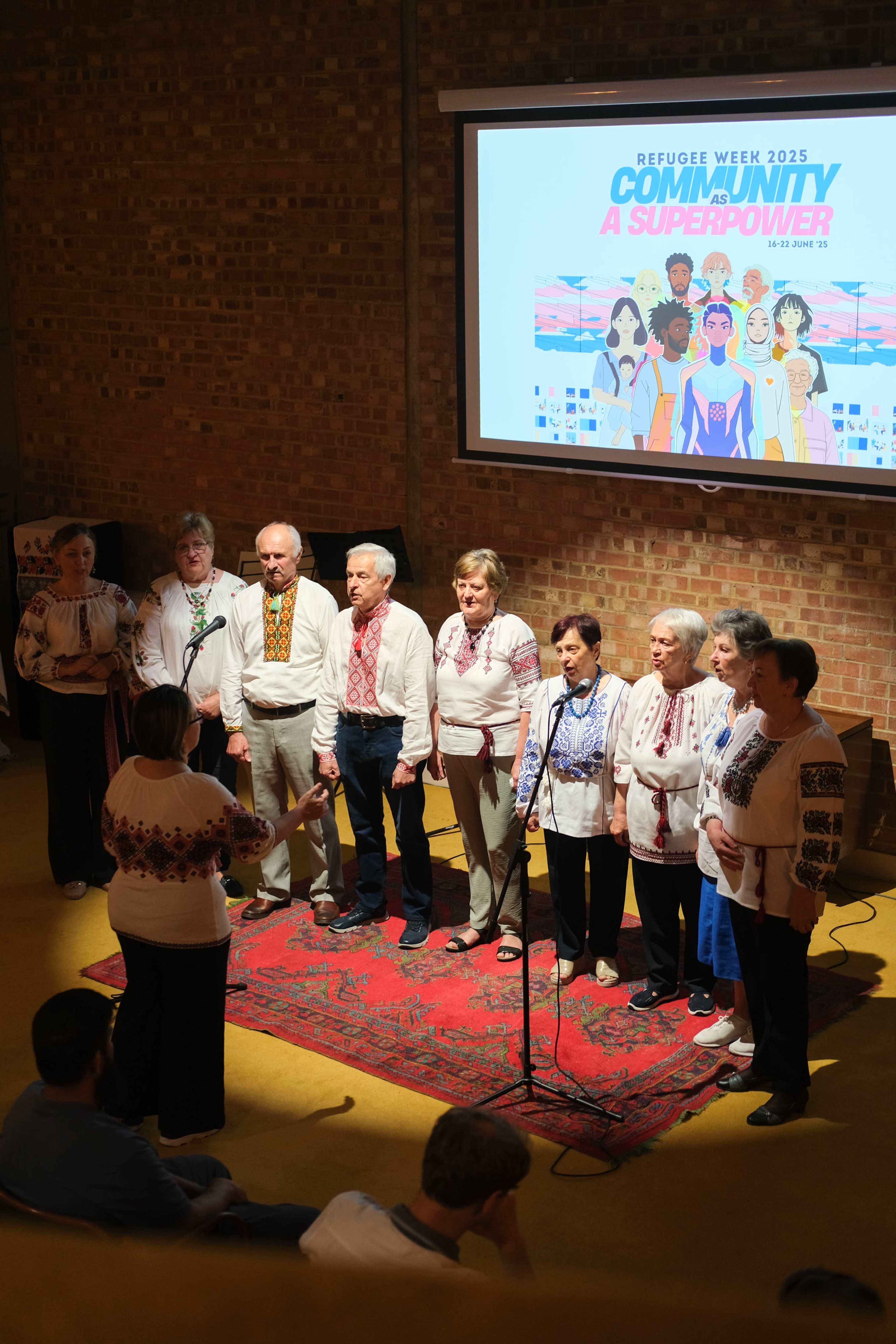 A group of people wearing traditional embroidered shirts stands on a stage, singing while a banner for Refugee Week 2025 is displayed in the background.