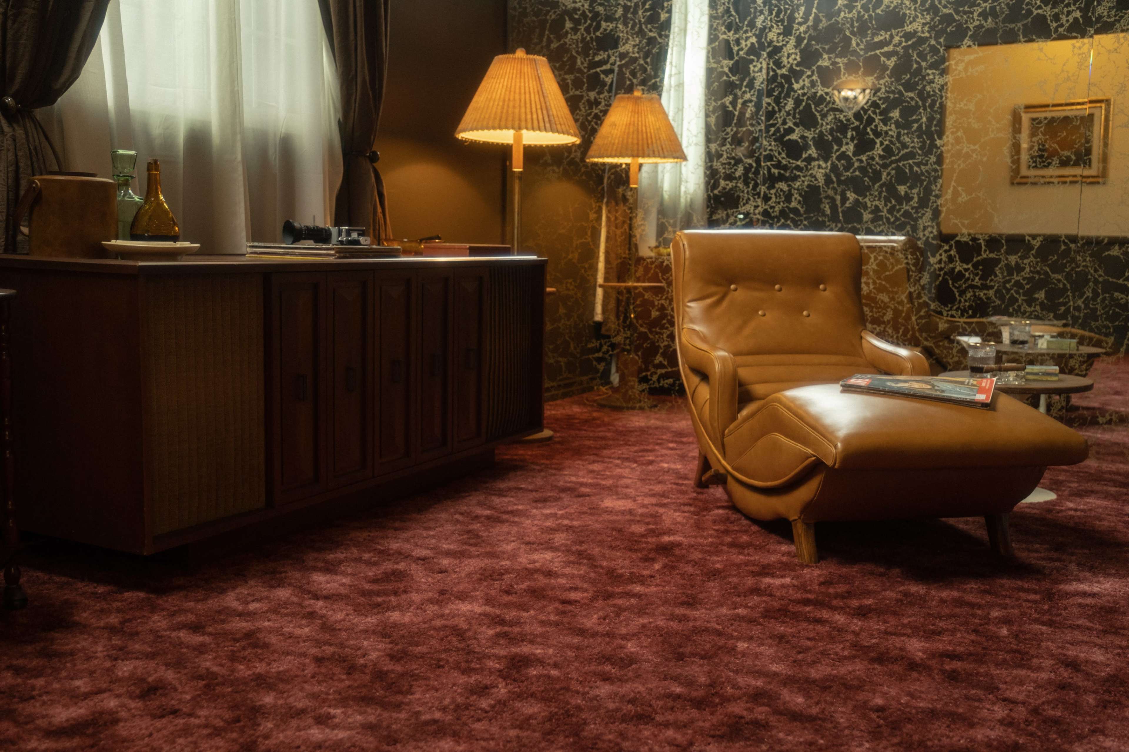 The scene features a cozy lounge area with a brown leather chaise lounge, a wooden cabinet, and dim lighting from two lamps against a textured wall.