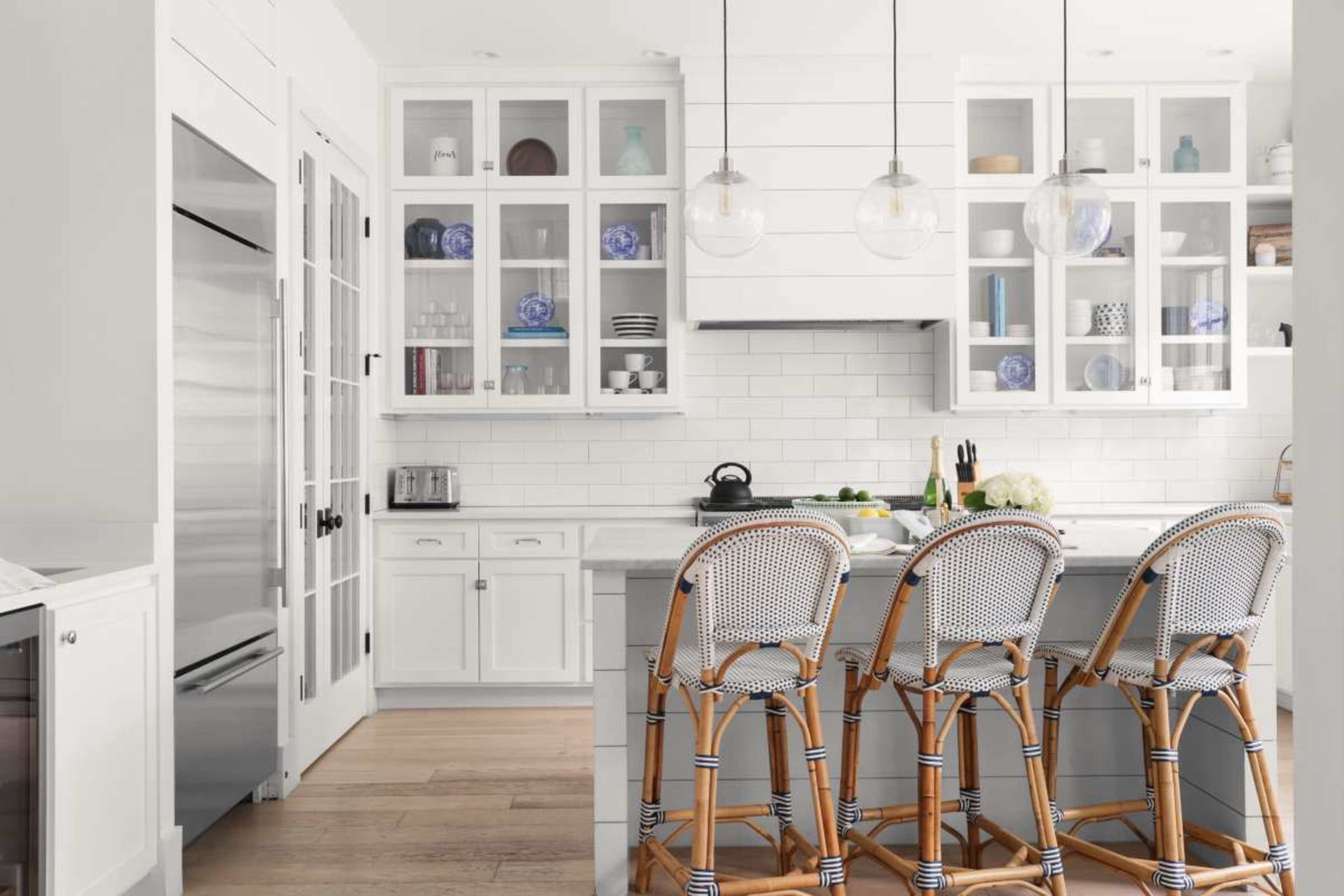 The image depicts a bright kitchen featuring white cabinetry, a large island with bar stools, and glass-fronted cabinets displaying decorative dishware.
