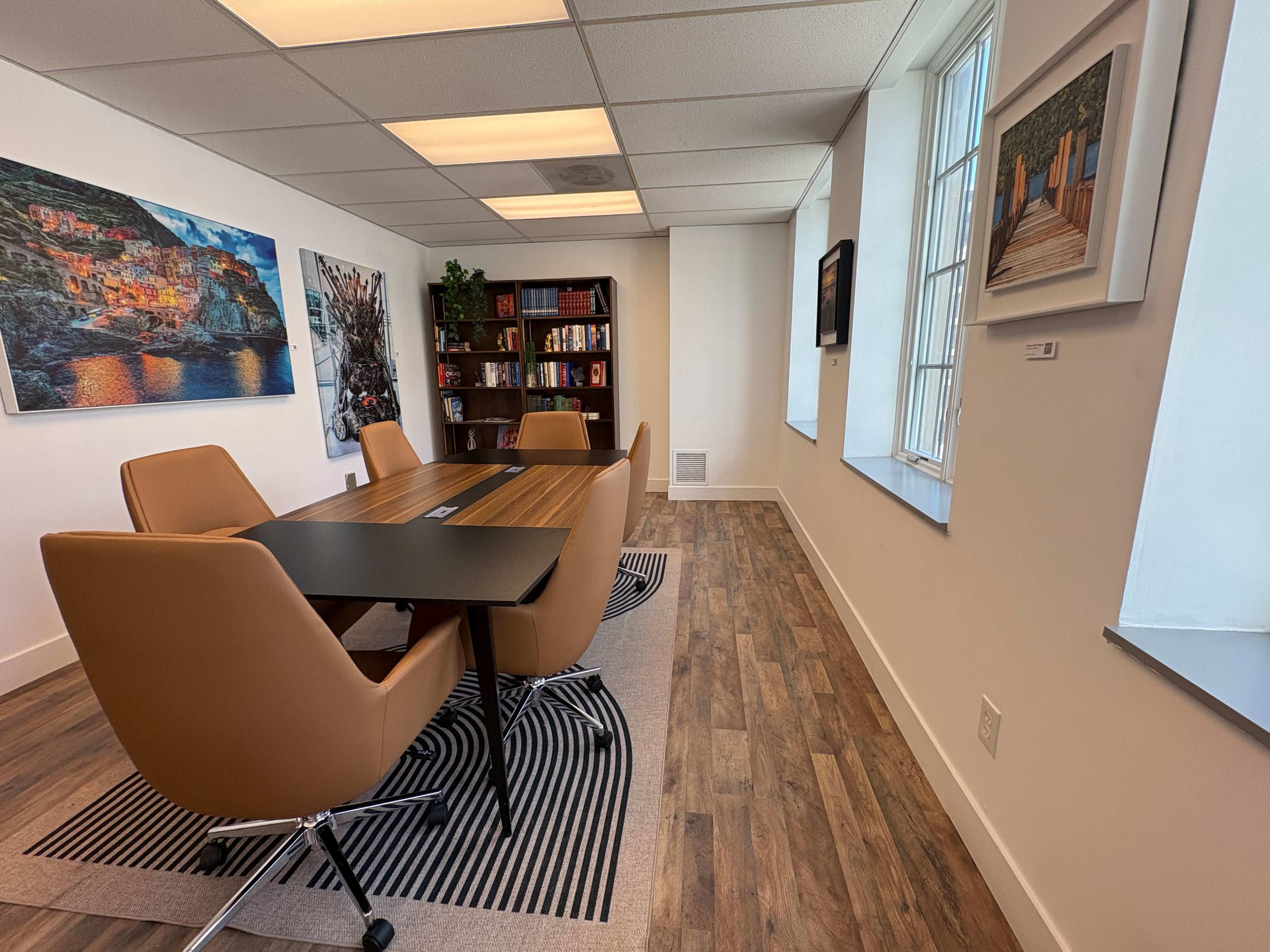 A modern conference room features a large wooden table surrounded by brown leather chairs, with walls decorated by artwork and a bookshelf filled with books.