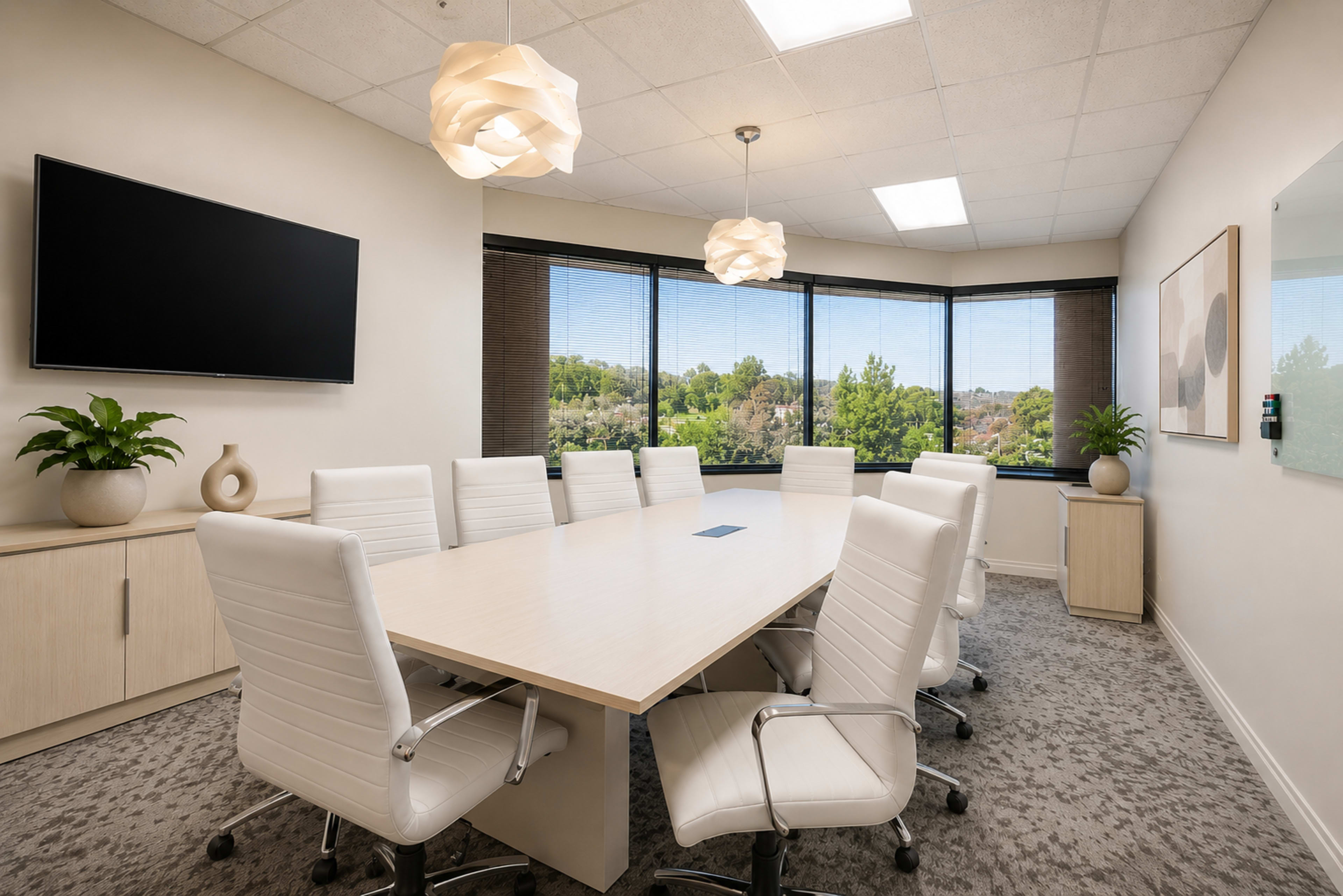 A modern conference room features a long table surrounded by white chairs and a large window showcasing a green view outside.