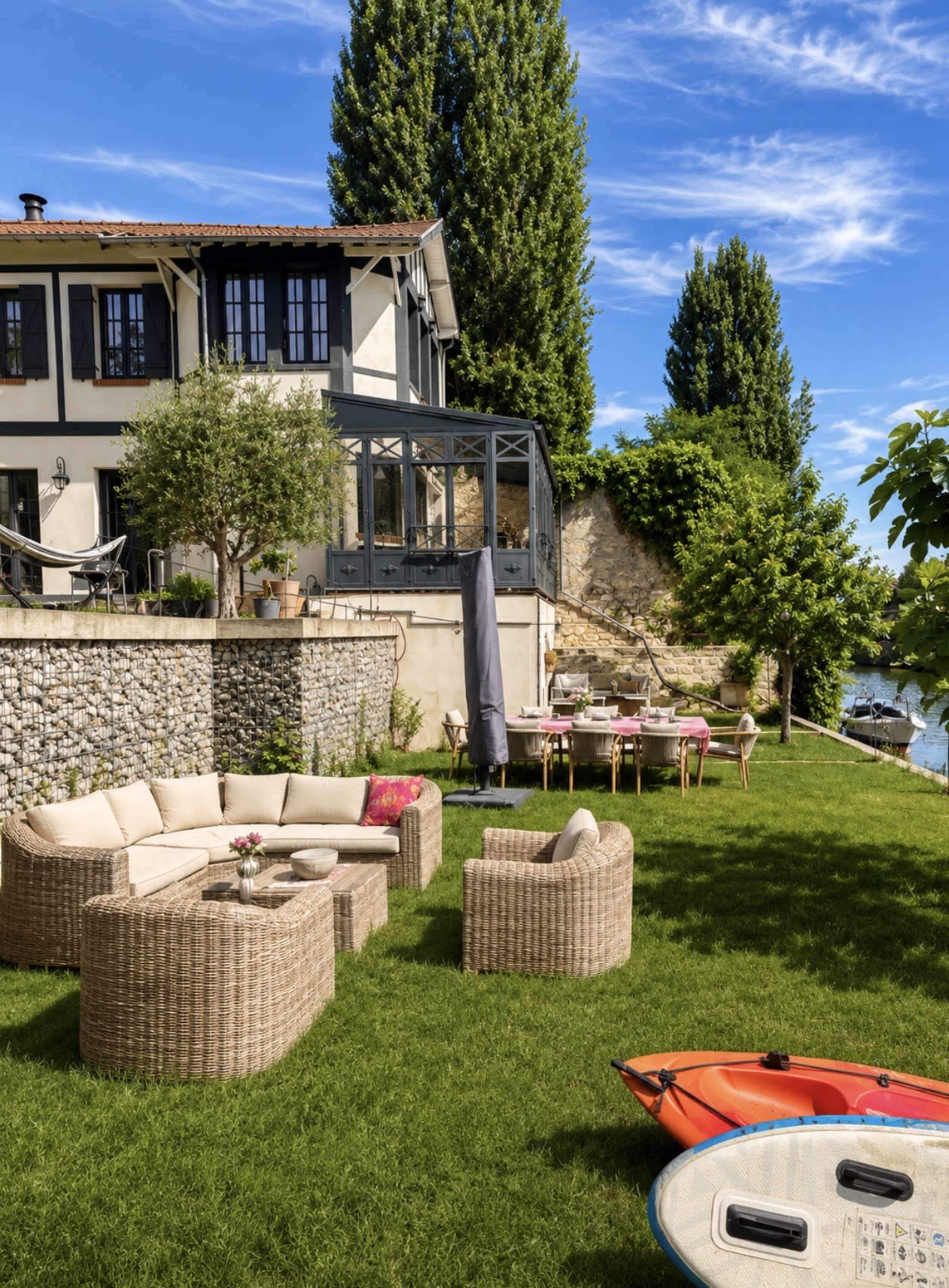 A spacious outdoor area features a modern home overlooking a body of water, with comfortable seating, a dining table, and kayaks nearby.
