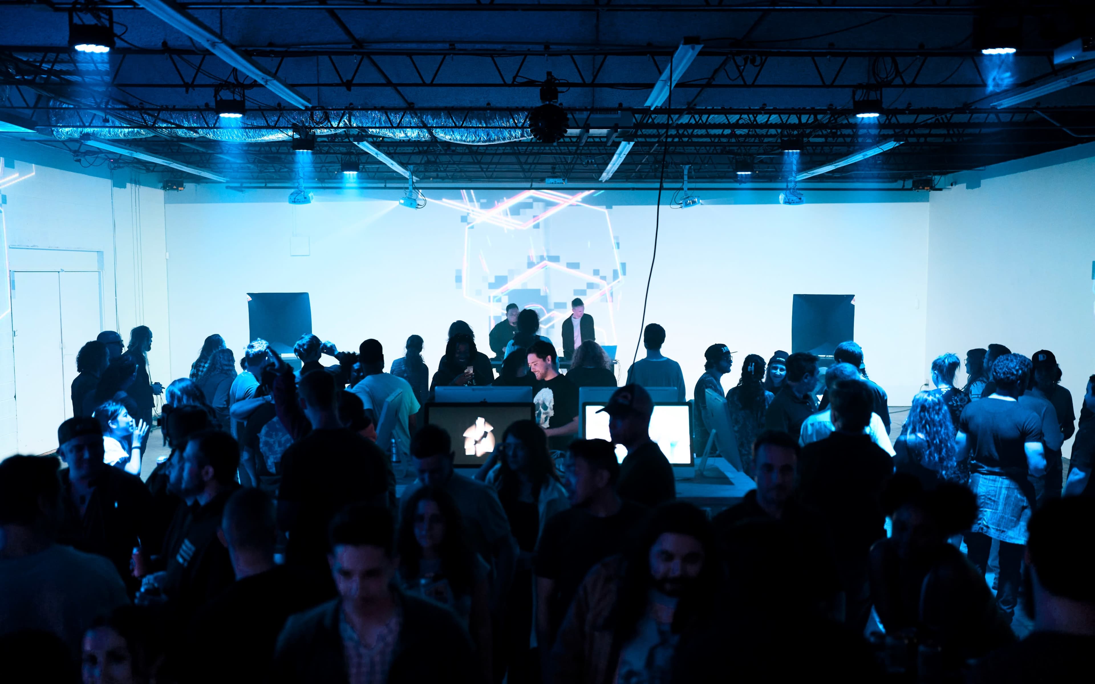 A crowd gathers in a dimly lit room with digital projections and DJ equipment setup, creating a lively atmosphere for a music event.