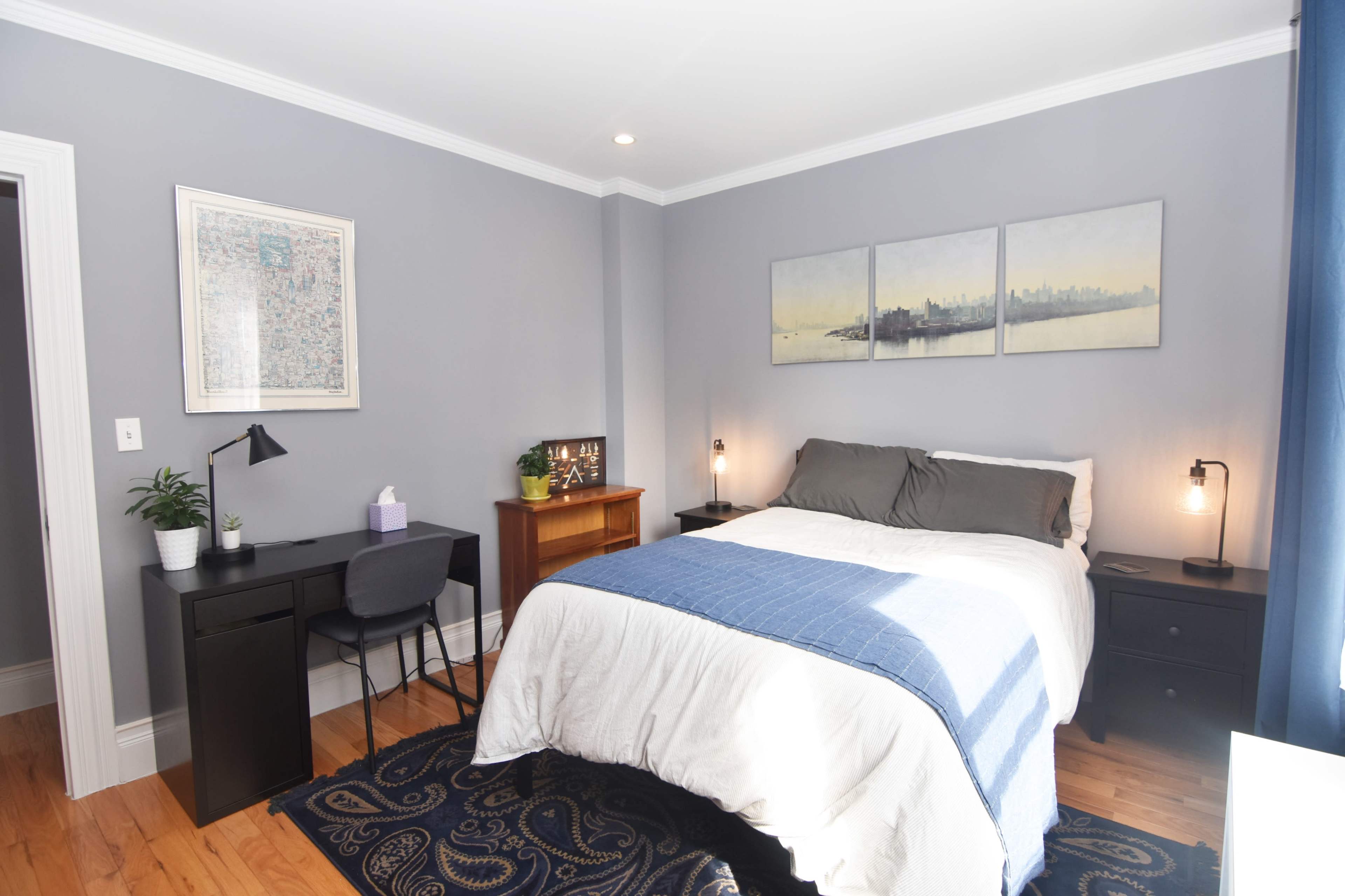 The image shows a neatly arranged bedroom featuring a bed with gray and white bedding, a desk with a chair, and artwork on the walls.