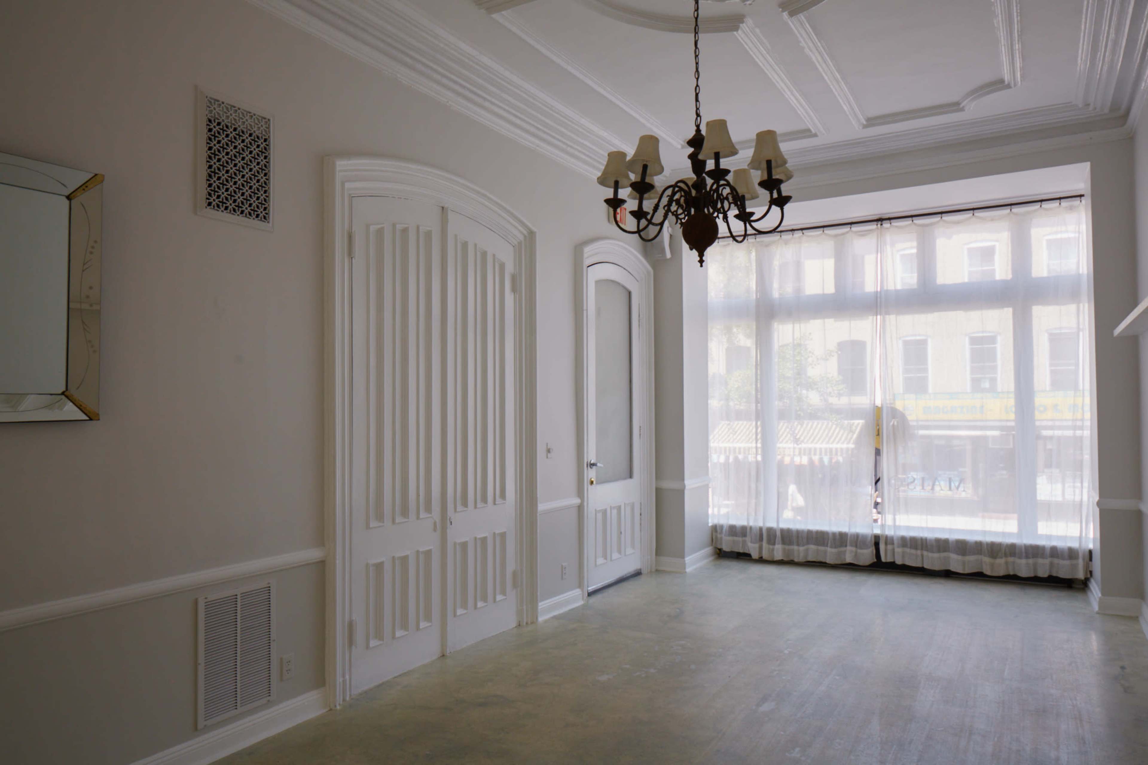 A spacious room with white walls, a chandelier, and large windows that overlook a street.