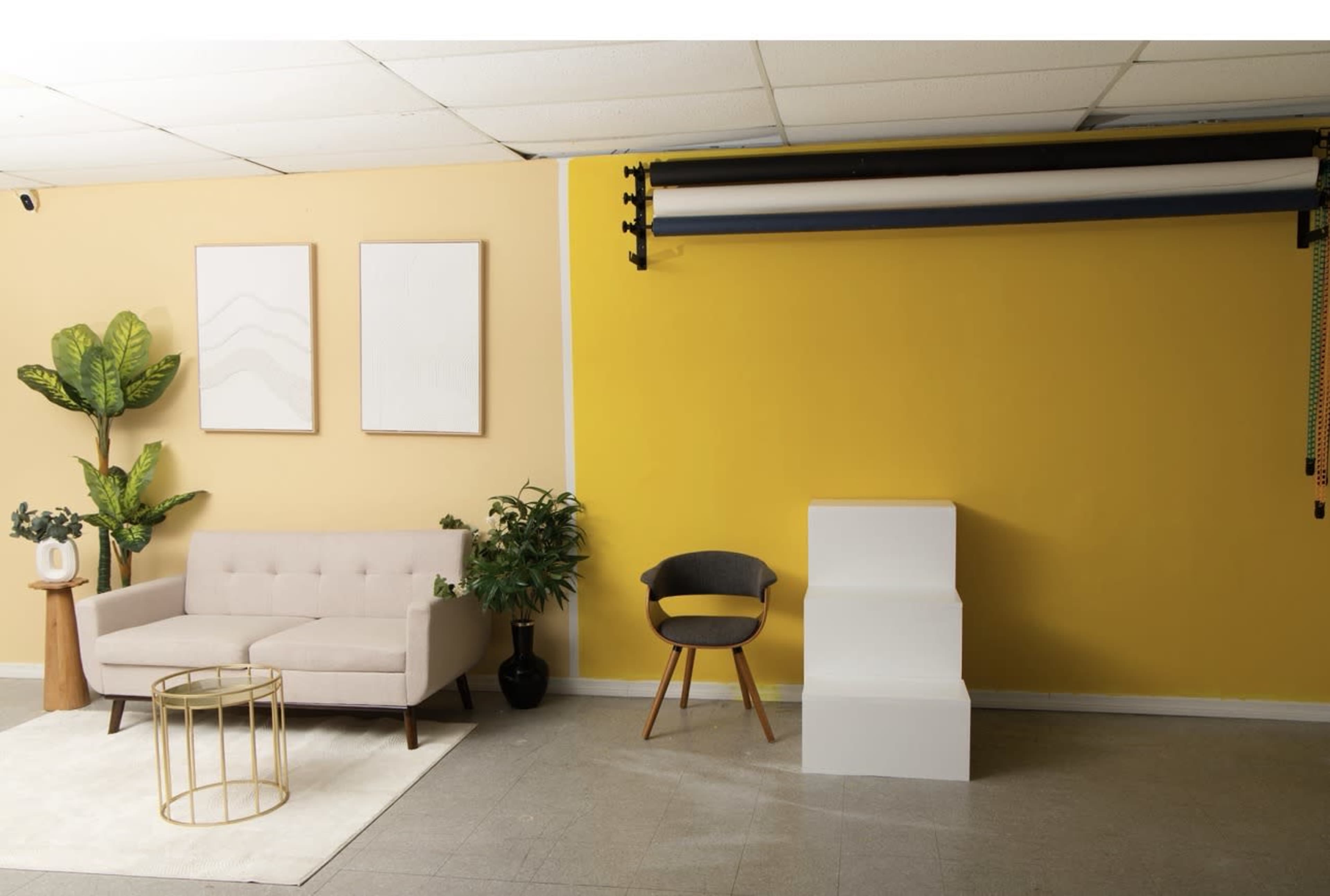 The image features a spacious room with a yellow accent wall, a beige sofa, a round side table, a plant, and a simple white display stand.