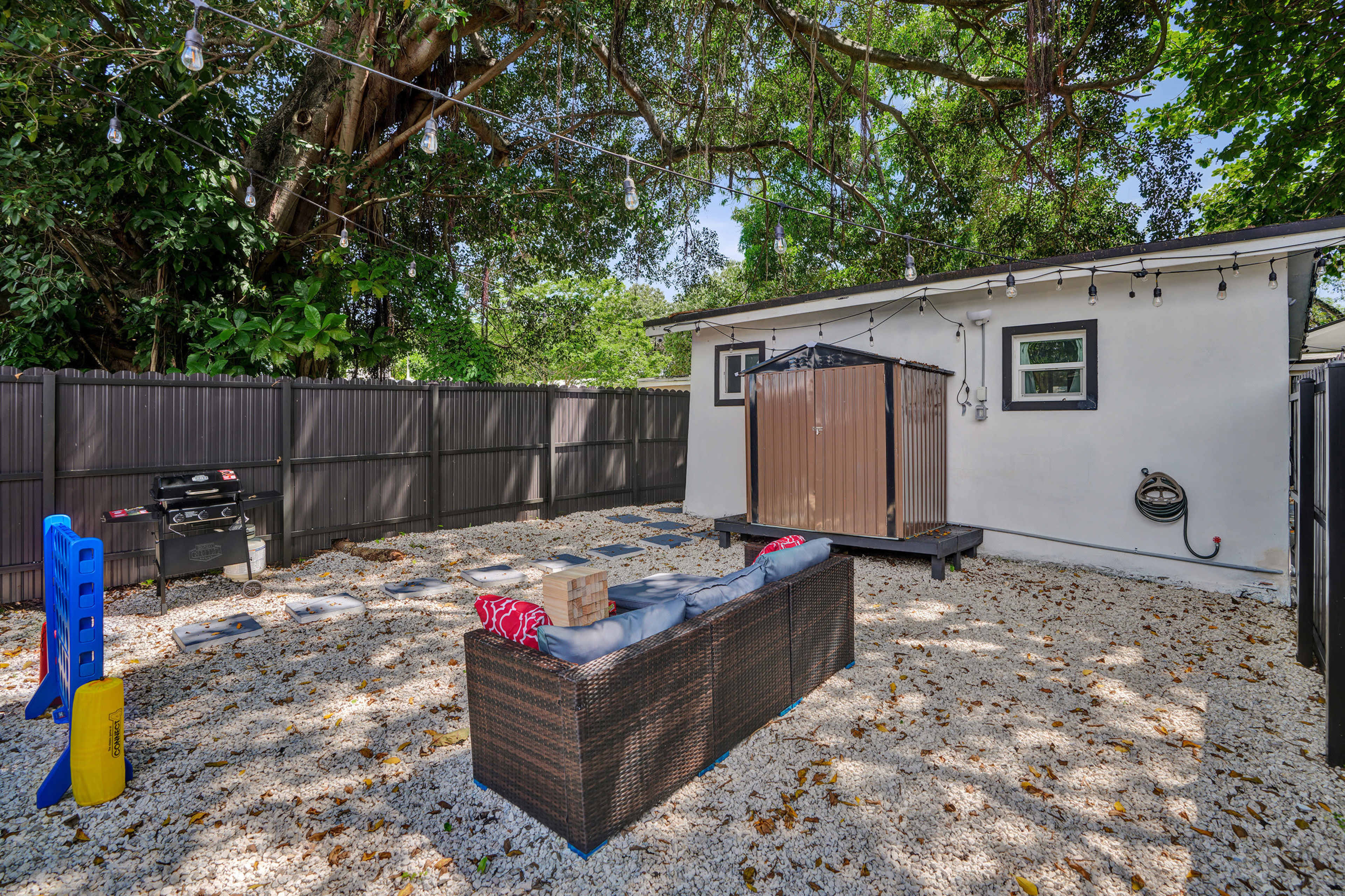 The image shows a fenced backyard with gravel flooring, a seating area topped with cushions, a grill, and a small shed or storage unit, all surrounded by greenery and string lights.