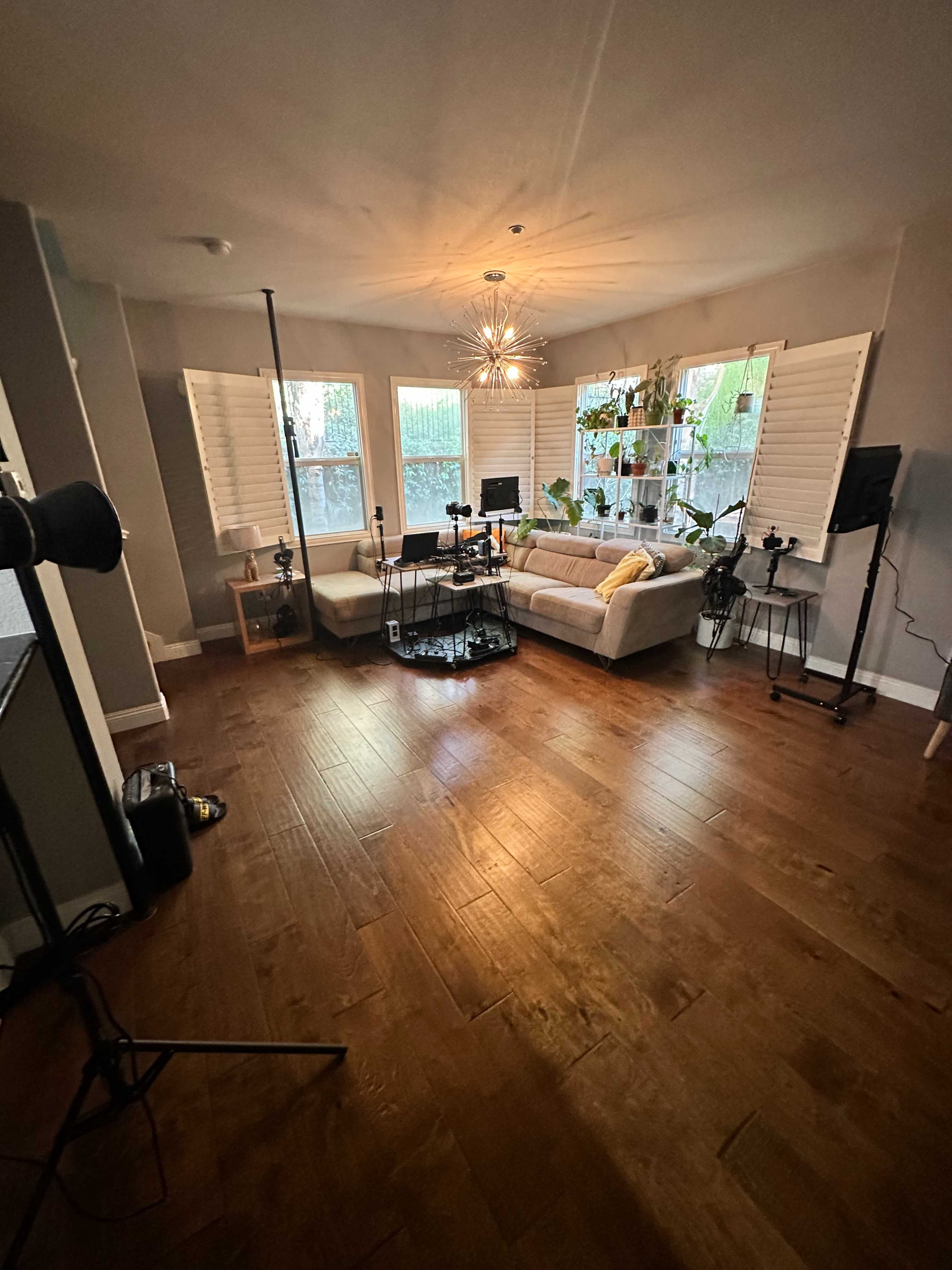 A well-lit living room with a sectional couch, plants in the windows, and a setup for recording gear.