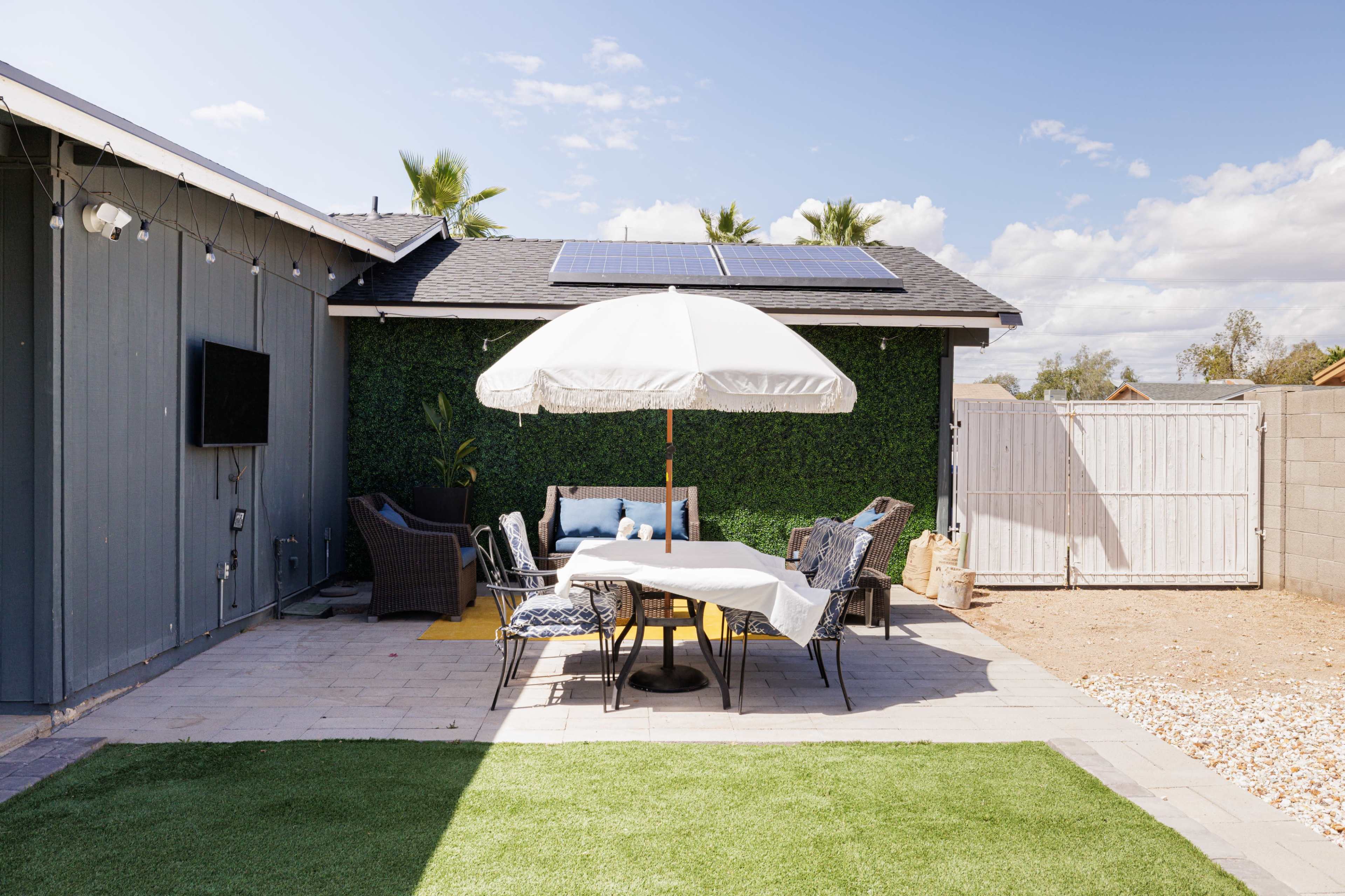 A patio area features a table with chairs under a large umbrella, surrounded by greenery and solar panels on the roof.