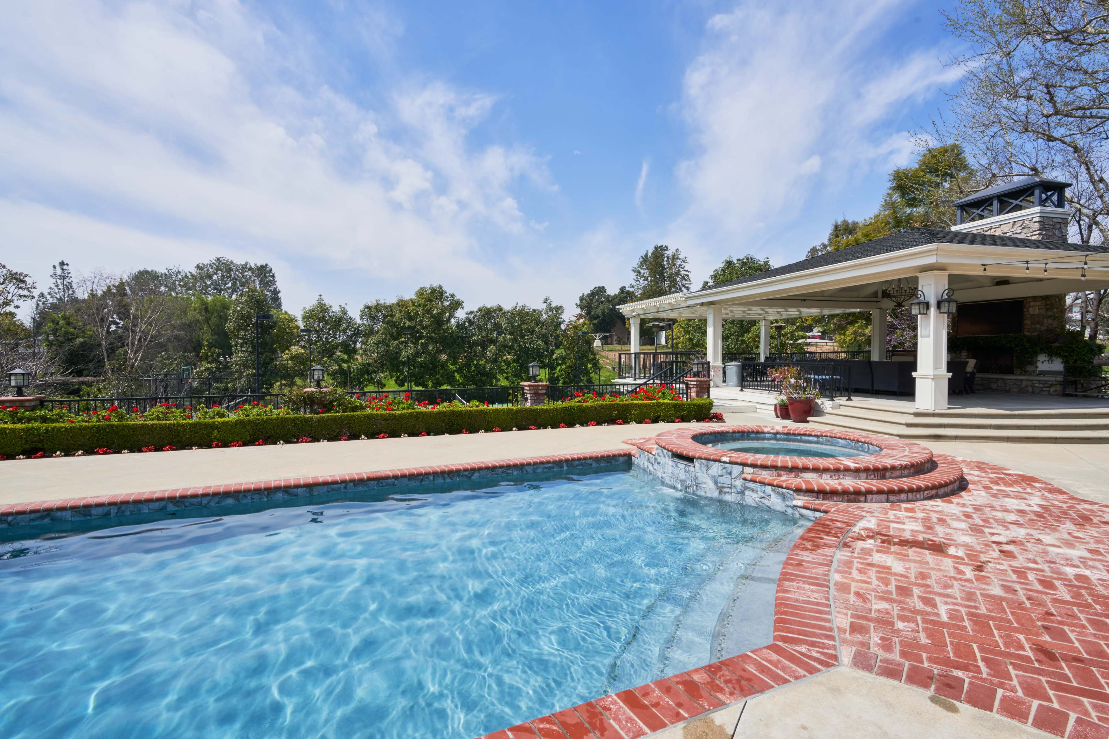 A swimming pool with a spa is surrounded by landscaped greenery and a covered pavilion.
