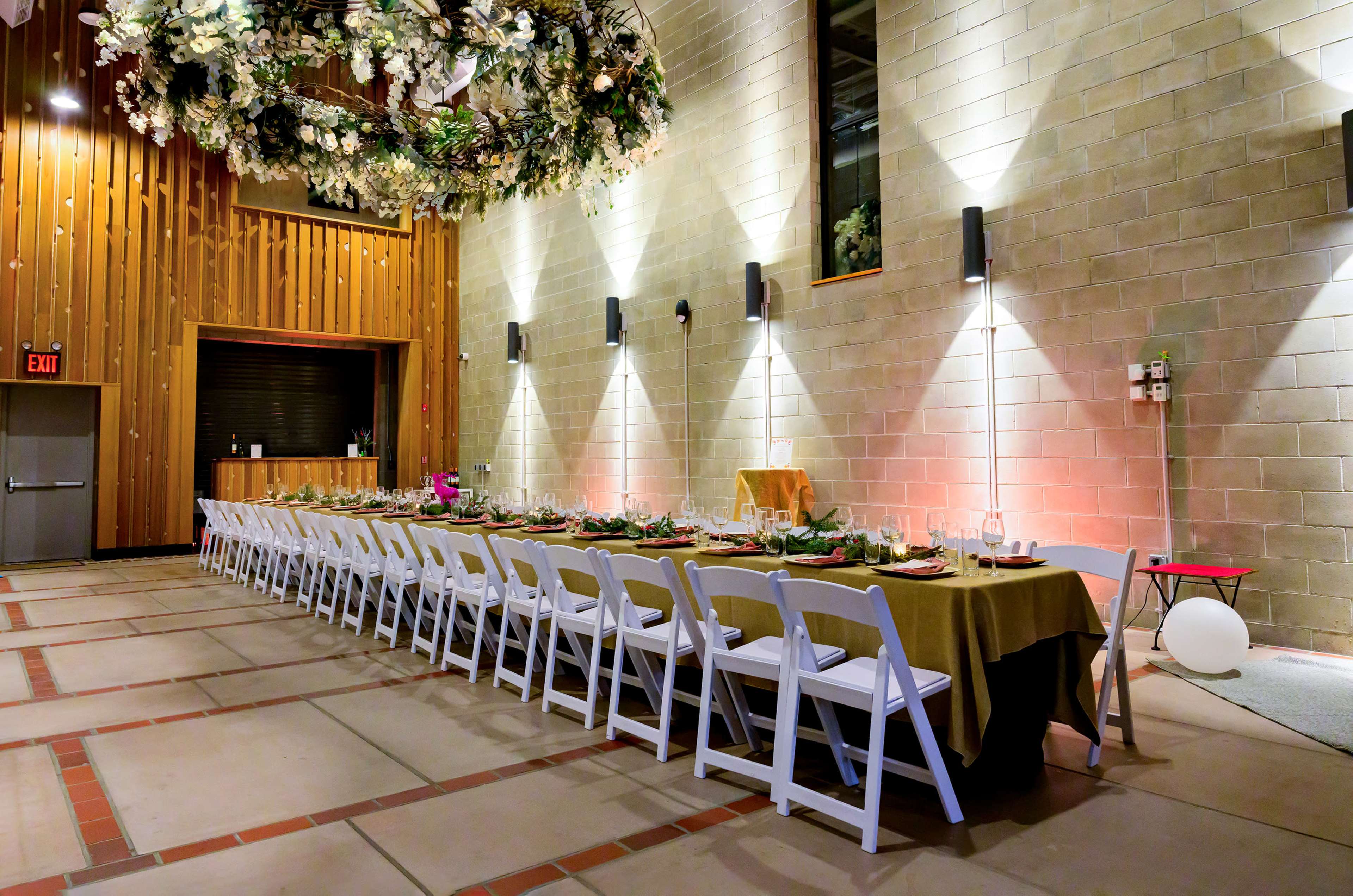 The image shows a long, elegantly set dining table in a spacious hall with wooden walls, decorated with floral arrangements and subtle lighting.
