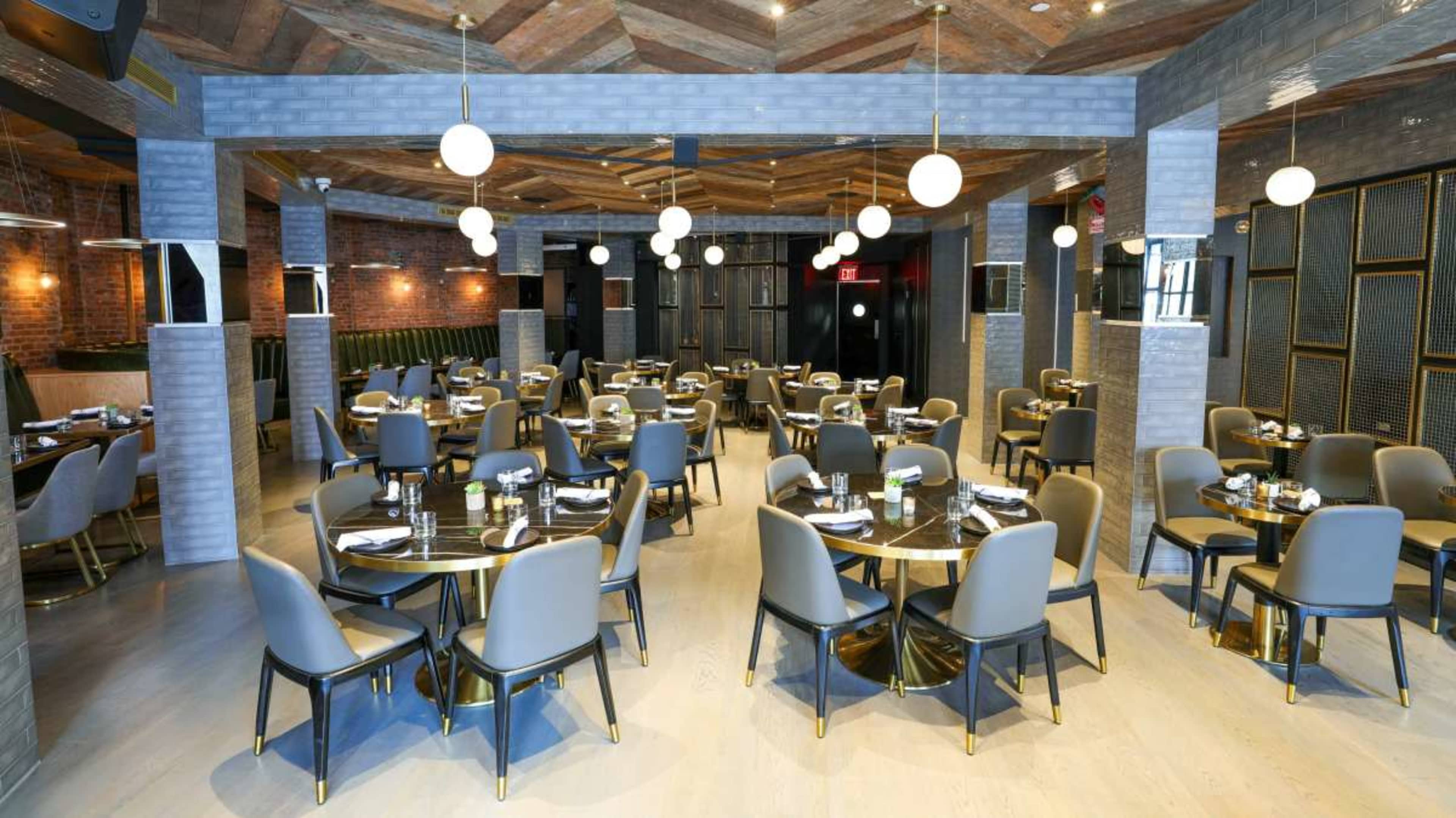 The image shows a modern restaurant interior with round tables and gray chairs arranged under pendant lights and a wooden ceiling.