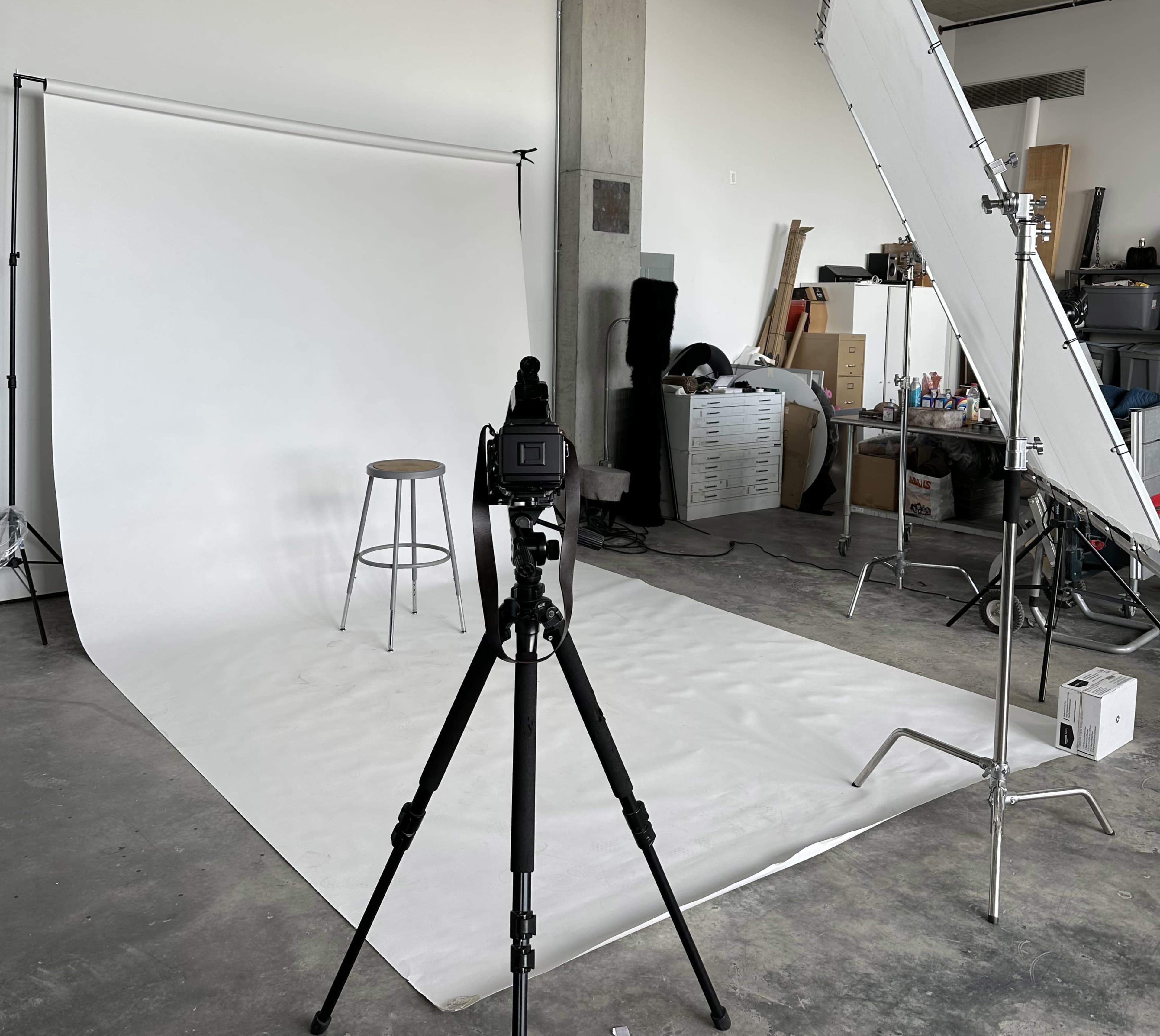 The image shows a photography setup in an industrial space, featuring a camera on a tripod facing a plain backdrop with a stool in front and a large reflector to the side.