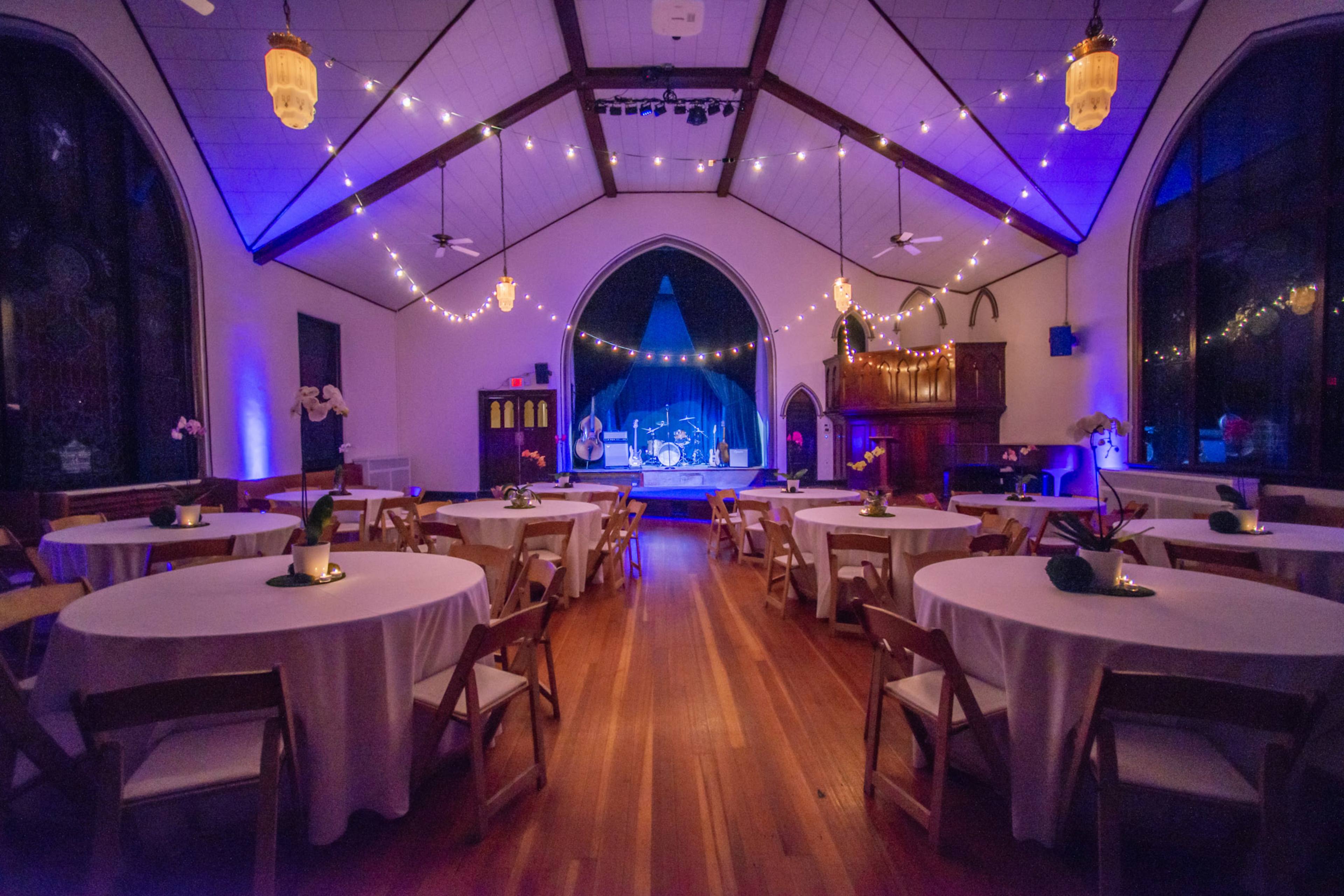 An elegantly decorated event space with round tables, wooden flooring, and a stage illuminated by soft blue and purple lighting.
