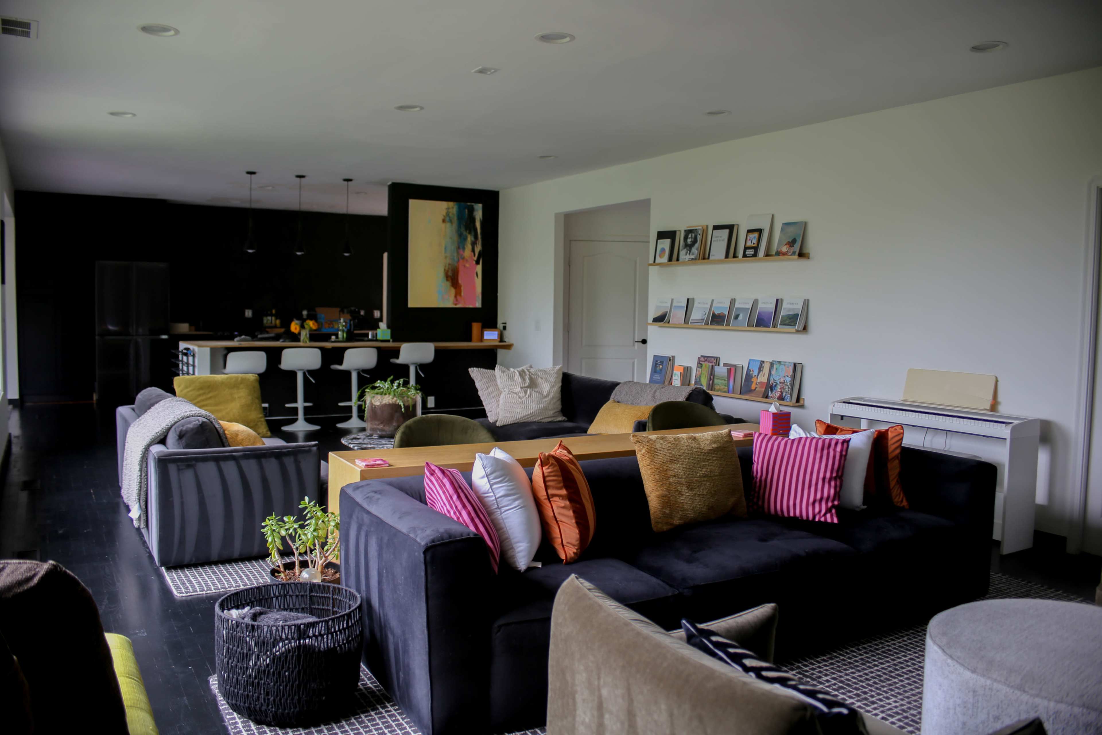 The image shows a modern living room with a combination of colorful sofas, a central coffee table, and a kitchen area in the background.