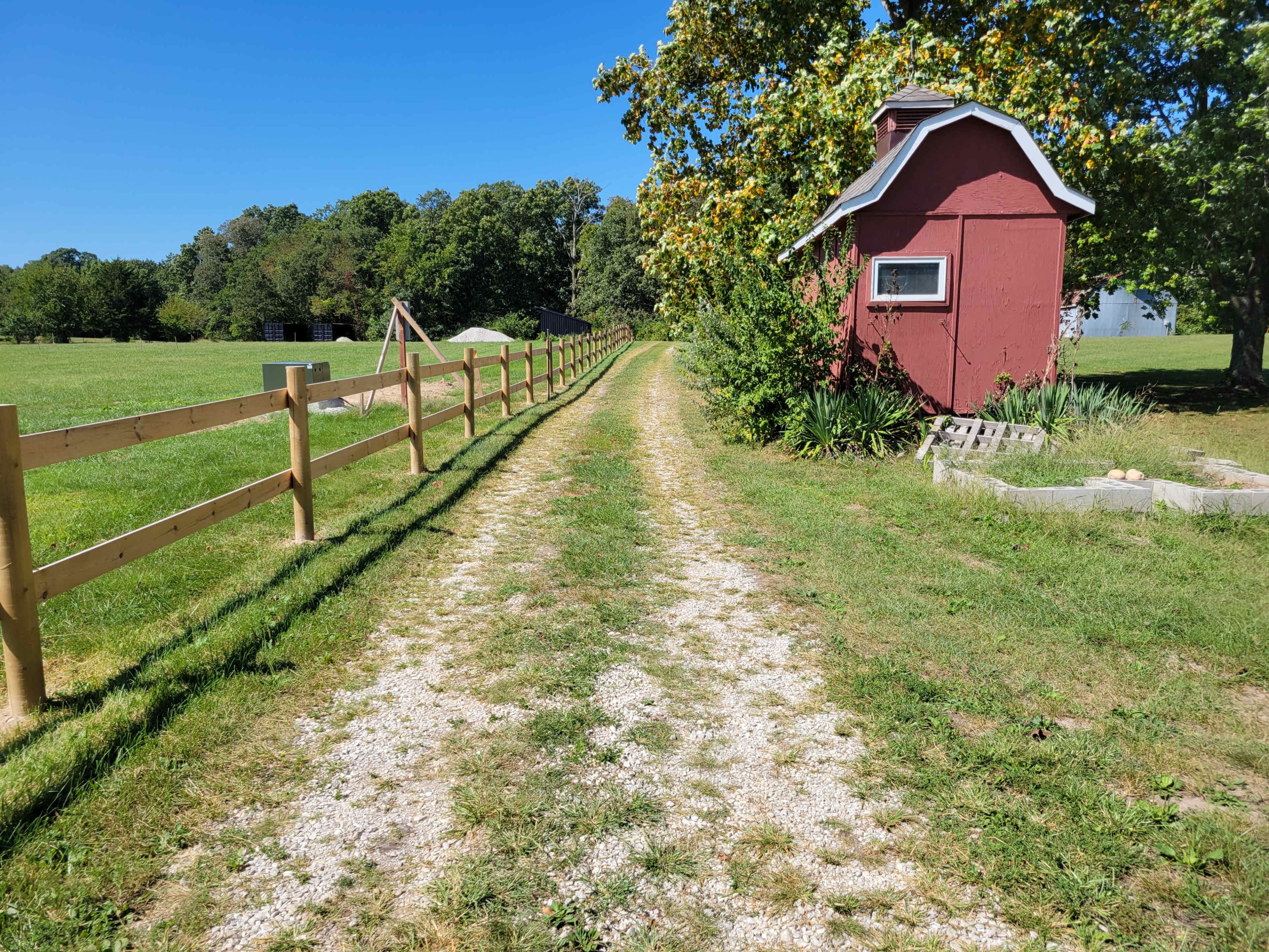 A gravel path leads through a fenced area beside a small red shed, with grassy fields and trees in the background.