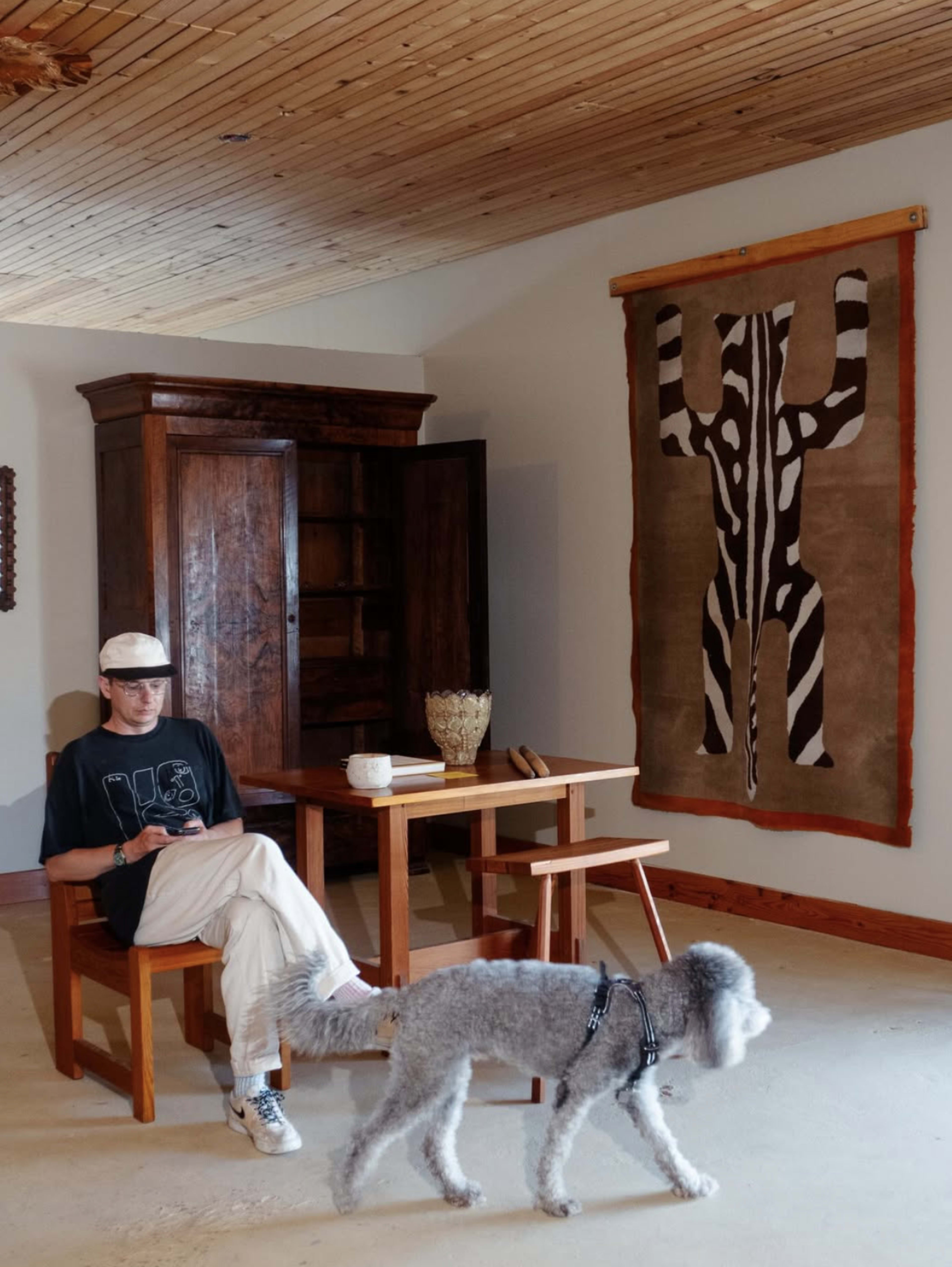 A person sitting on a chair beside a wooden table in a room with a large wall hanging and a dog walking nearby.
