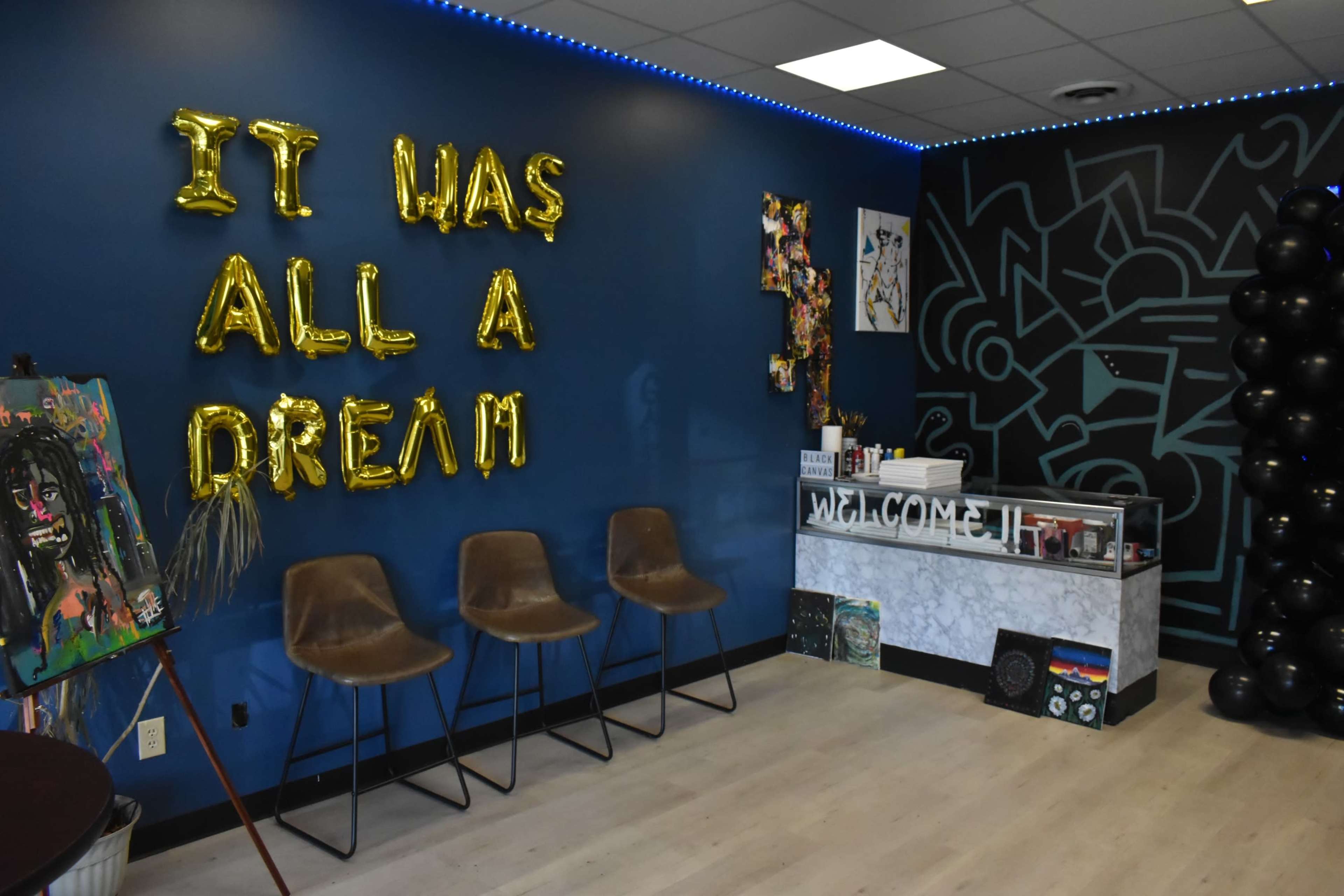 A modern reception area with gold balloon letters spelling "IT HAS ALL A DREAM" on a blue wall, accompanied by art pieces, chairs, and a reception desk.
