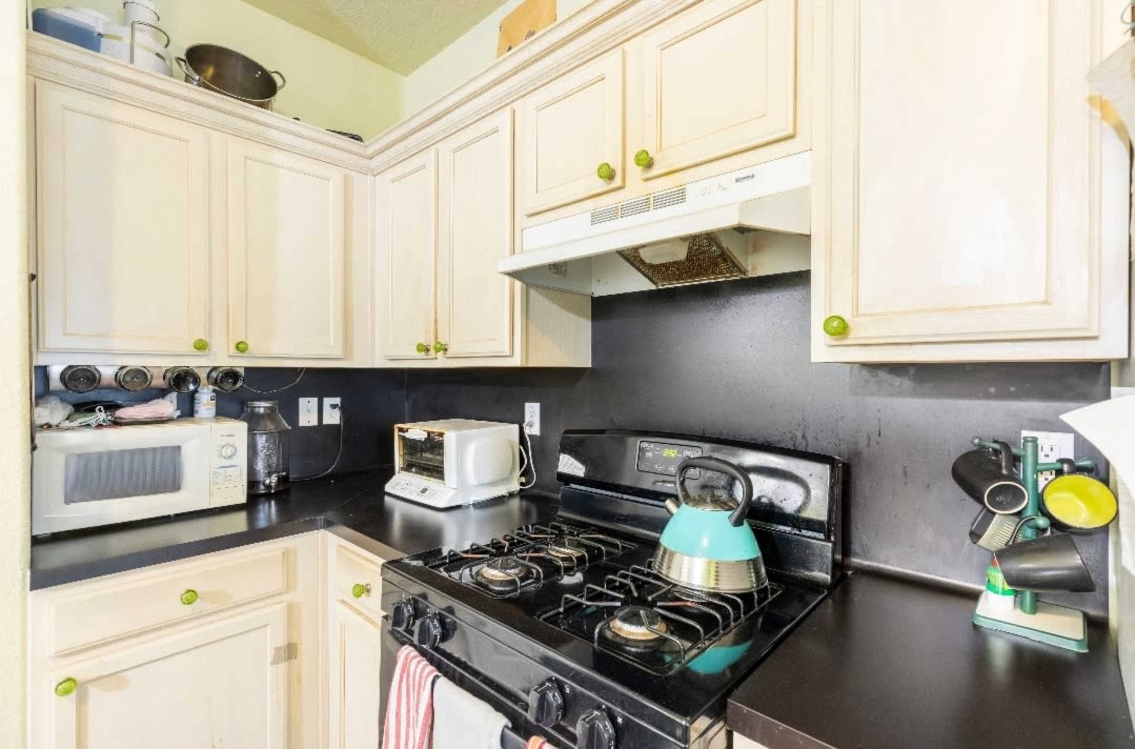 A compact kitchen with cream-colored cabinets, a black countertop, a gas stove with a kettle, and a microwave.