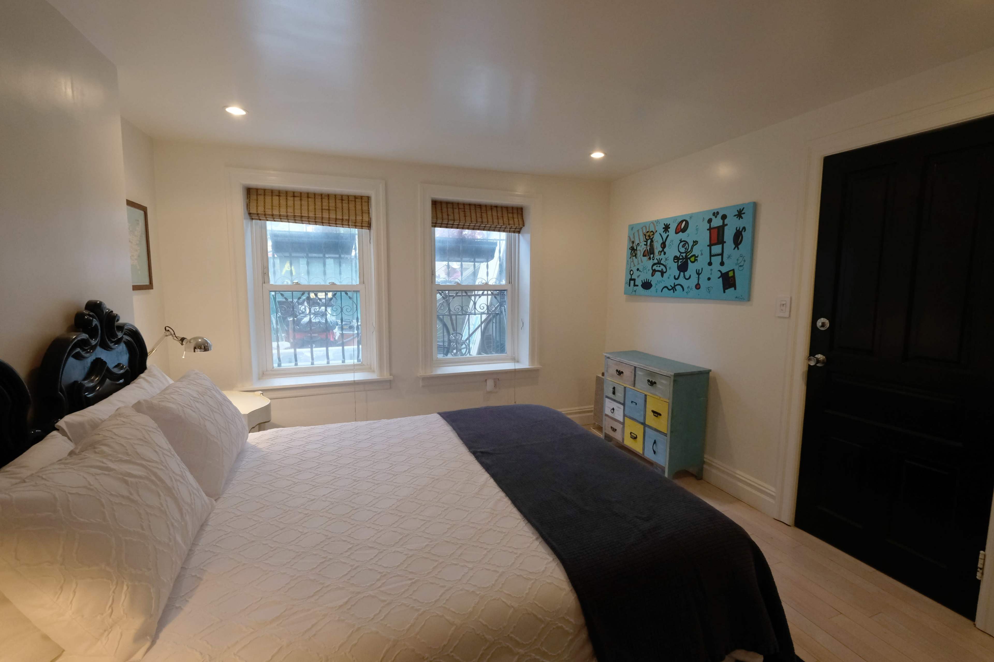 The image shows a well-lit bedroom with a bed, two windows, a colorful piece of wall art, and a small dresser against one wall.